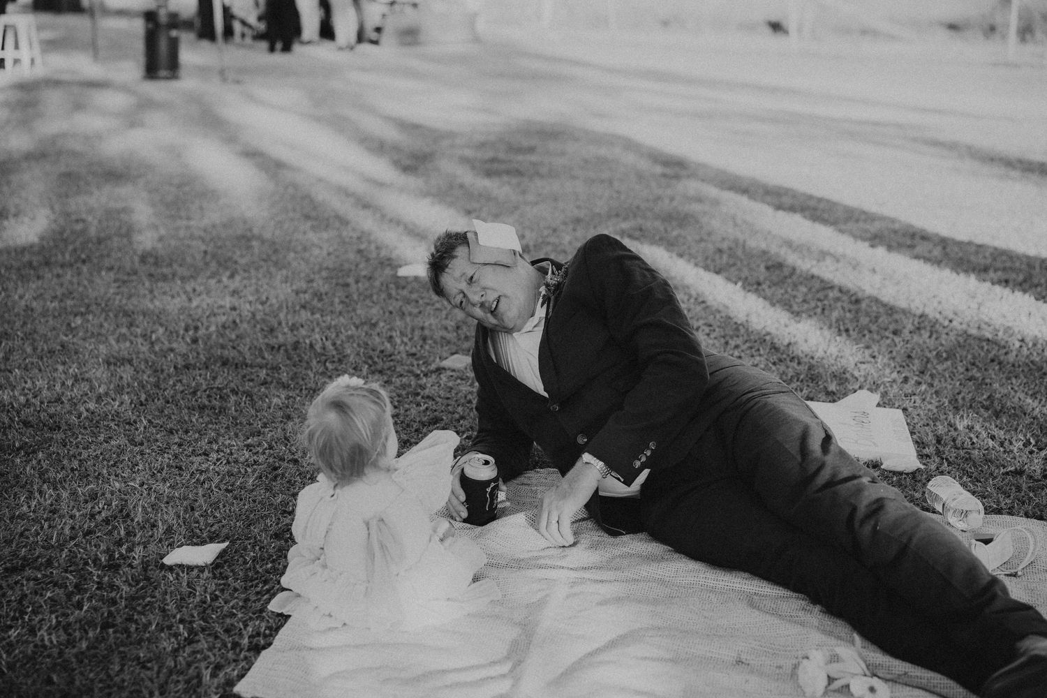 Man in a suit lies on a blanket on green grass interacting with a small child at a wedding reception.