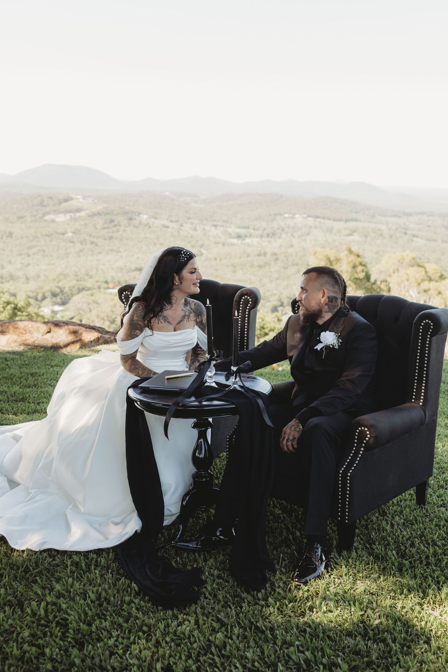 Newlywed couple relaxing together on black leather chairs on a grassy hillside.