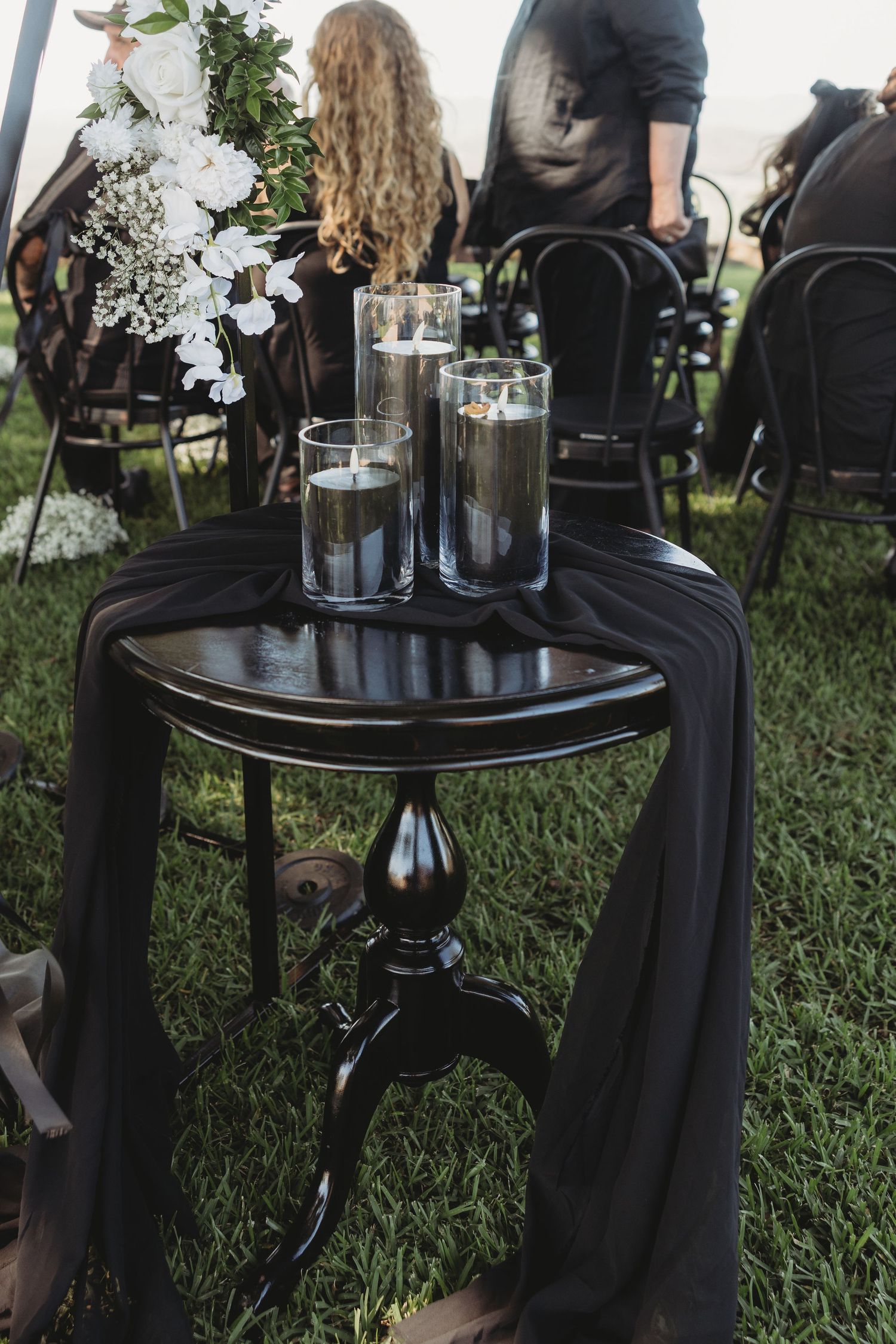 Round black side tables with floral arrangements sit on grass at an outdoor event setting.