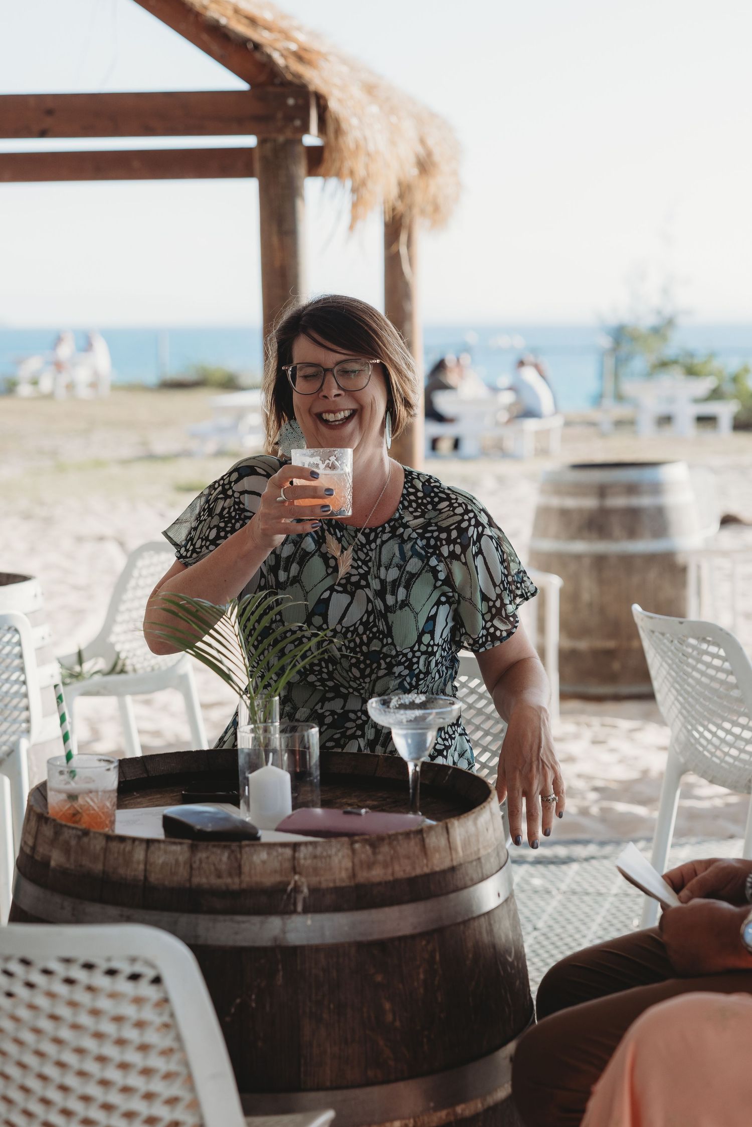 Person enjoying drinks at outdoor beach bar with wooden barrel tables.