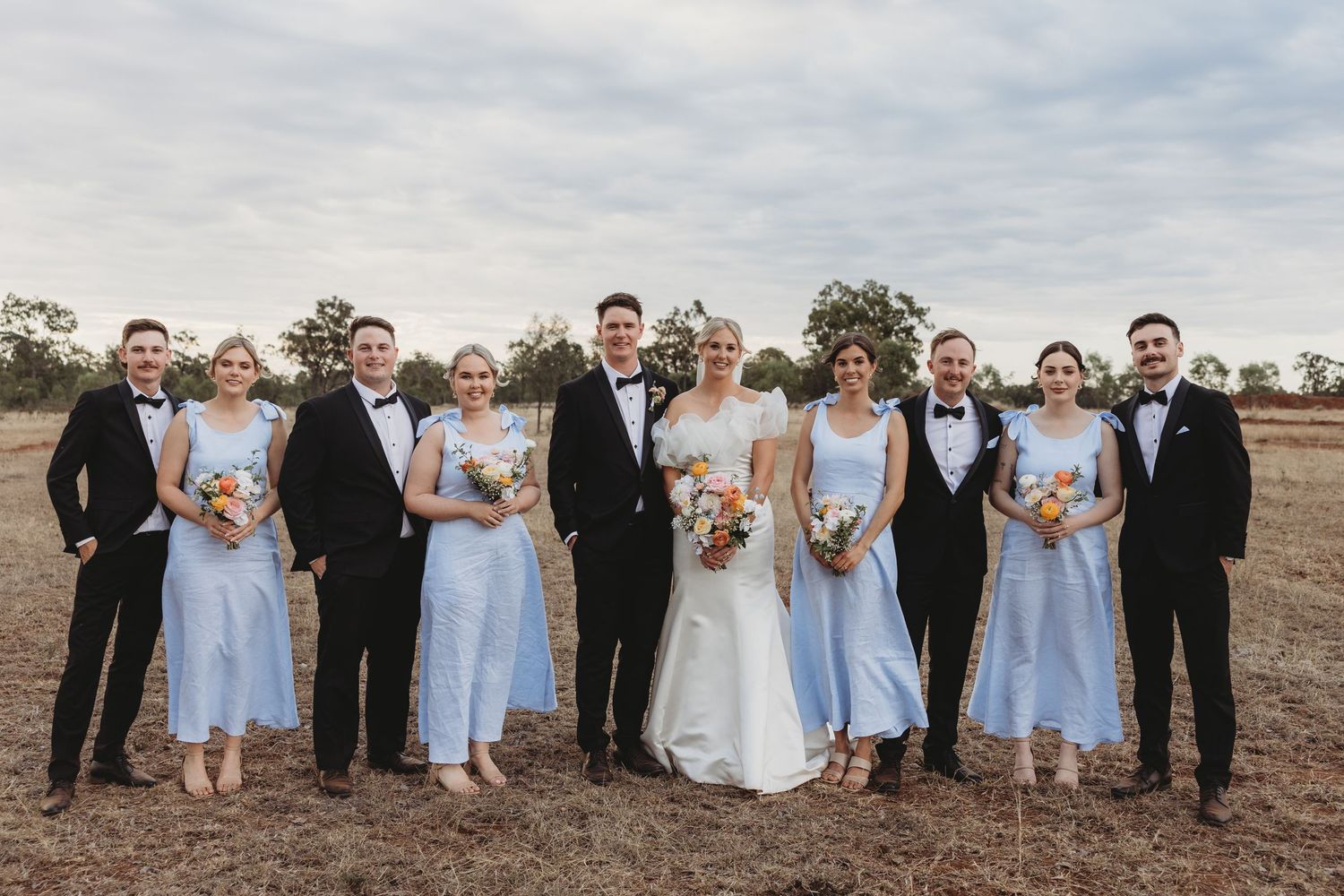 Wedding party in light blue dresses and black tuxedos poses together in an outdoor field setting.