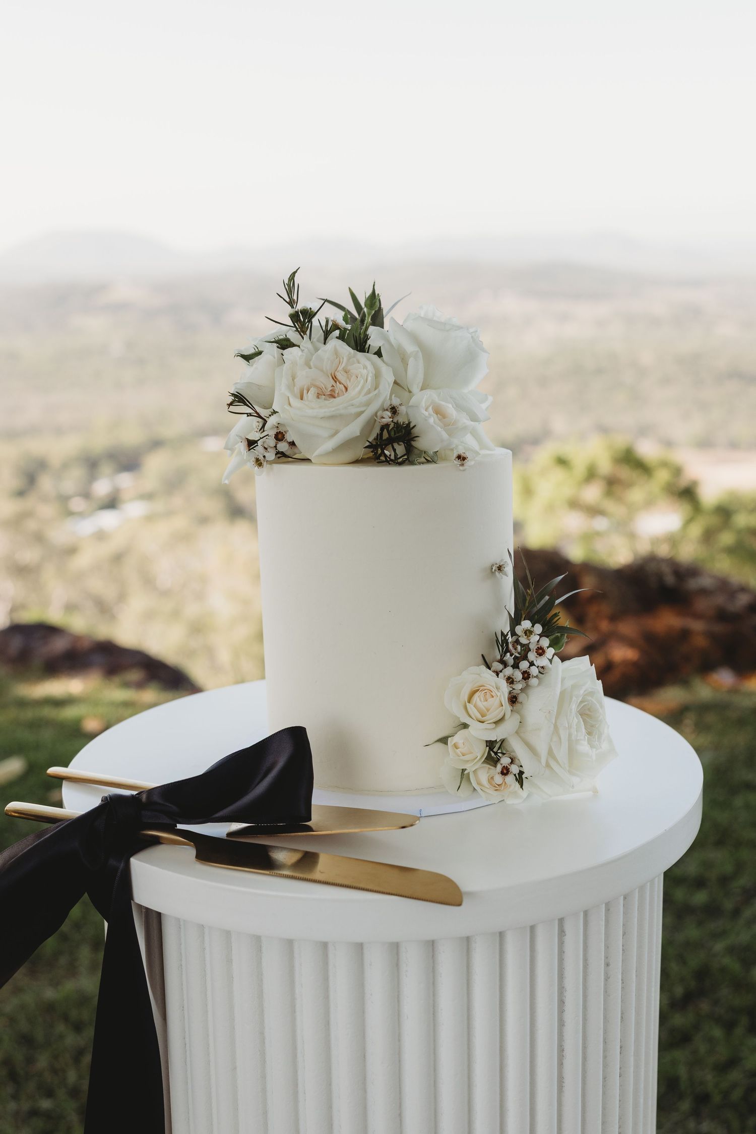 Modern white wedding cake with floral accents displayed on a ribbed column stand against a scenic backdrop.