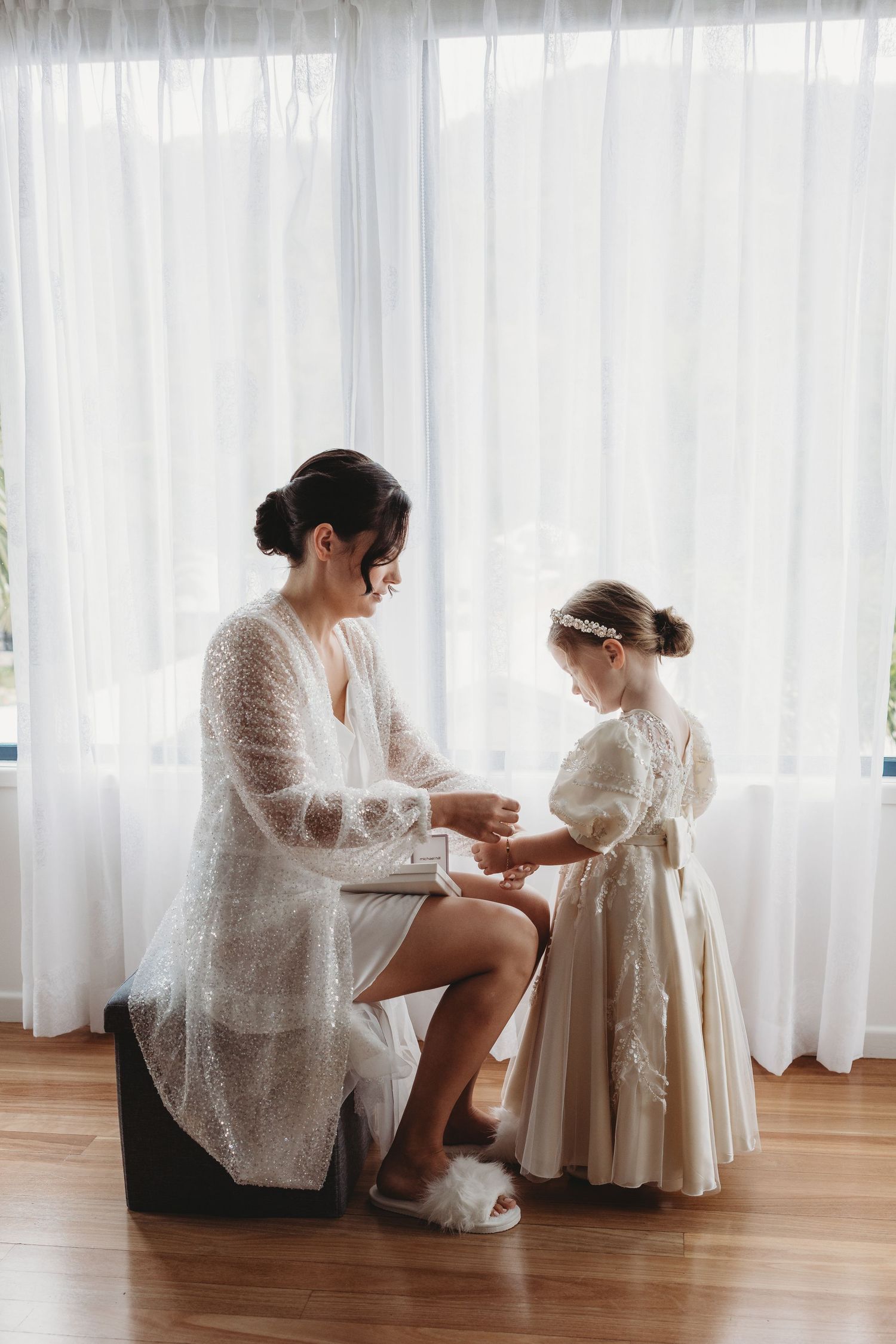 A tender moment between two people in white lace dresses by a window with sheer curtains.