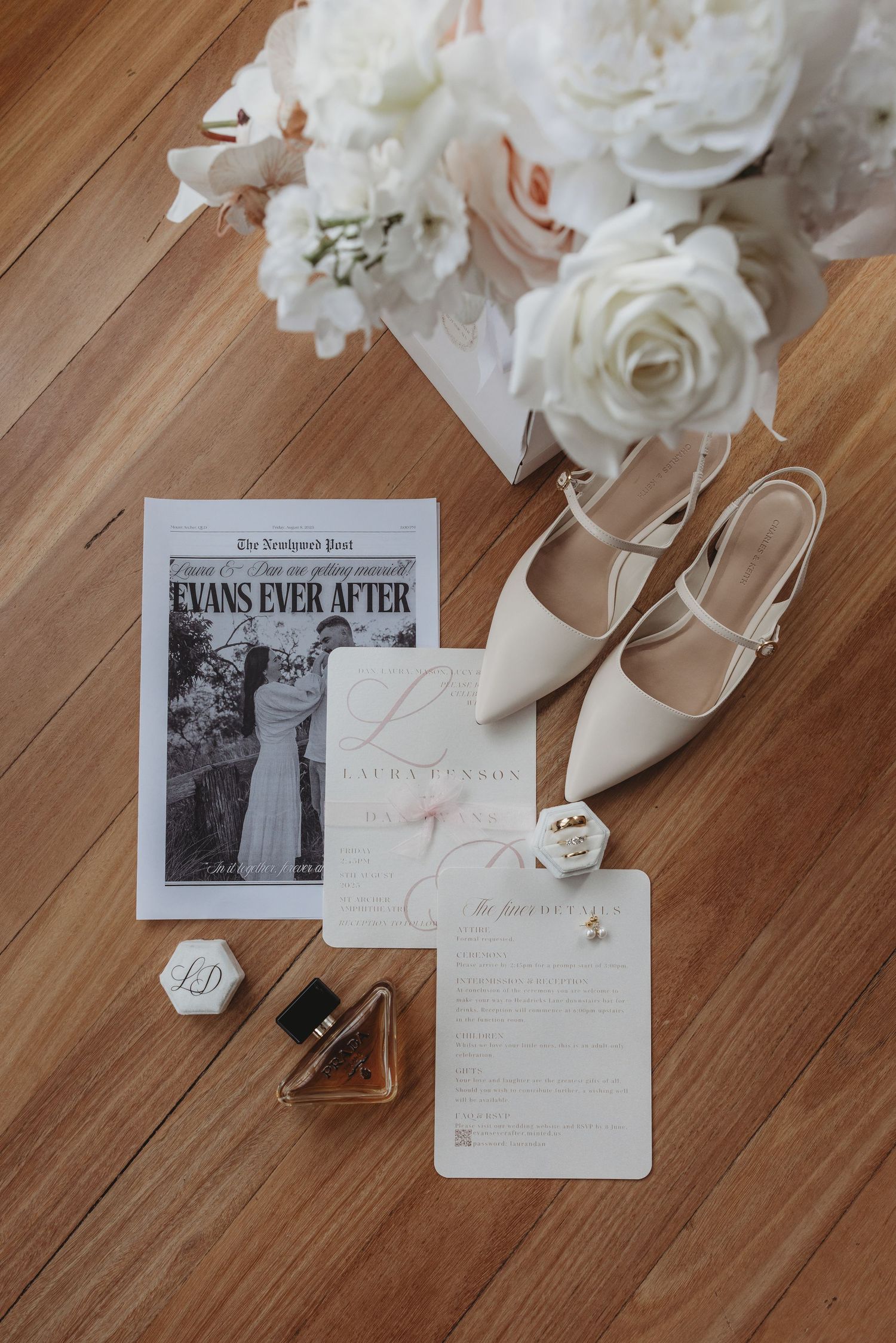 Wedding details flatlay with white flowers, shoes, and invitation cards on wooden floor.