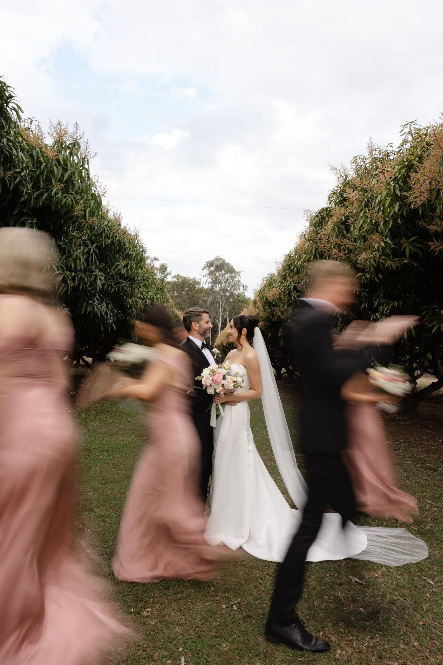 Bride and groom share a romantic moment as blurred wedding party moves around them in a lush garden setting.