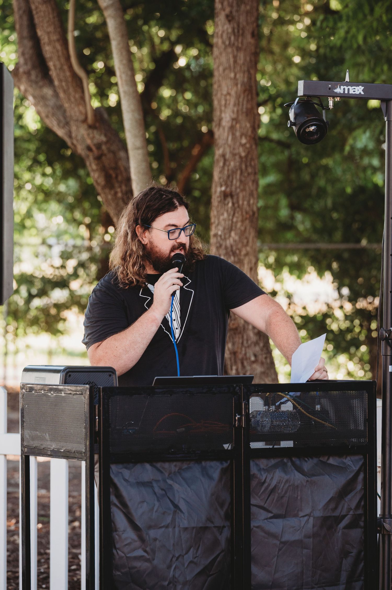 DJ equipment and sound booth setup under a covered outdoor venue with trees in the background.