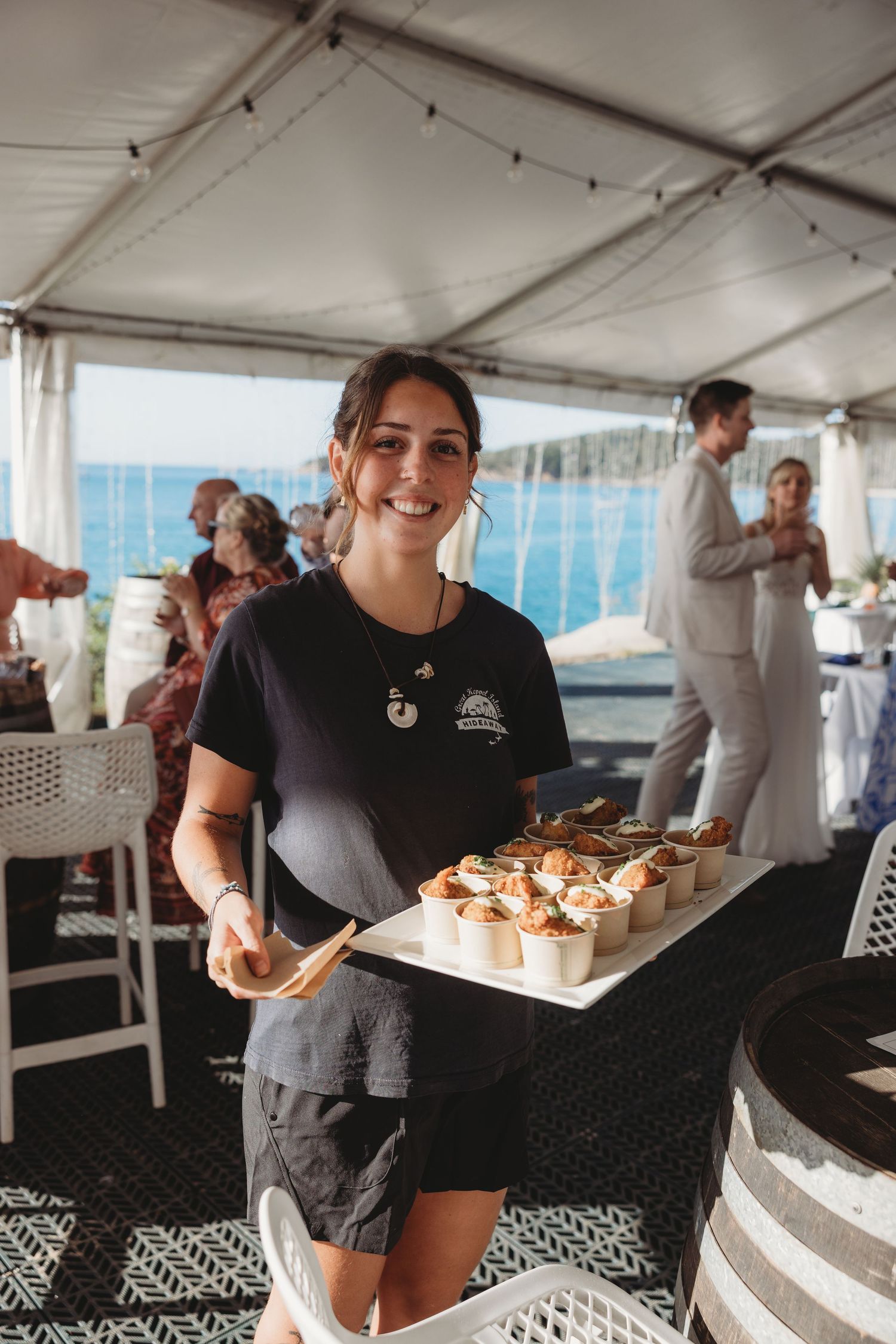 Catering staff serves appetizers under white tent at beach reception.