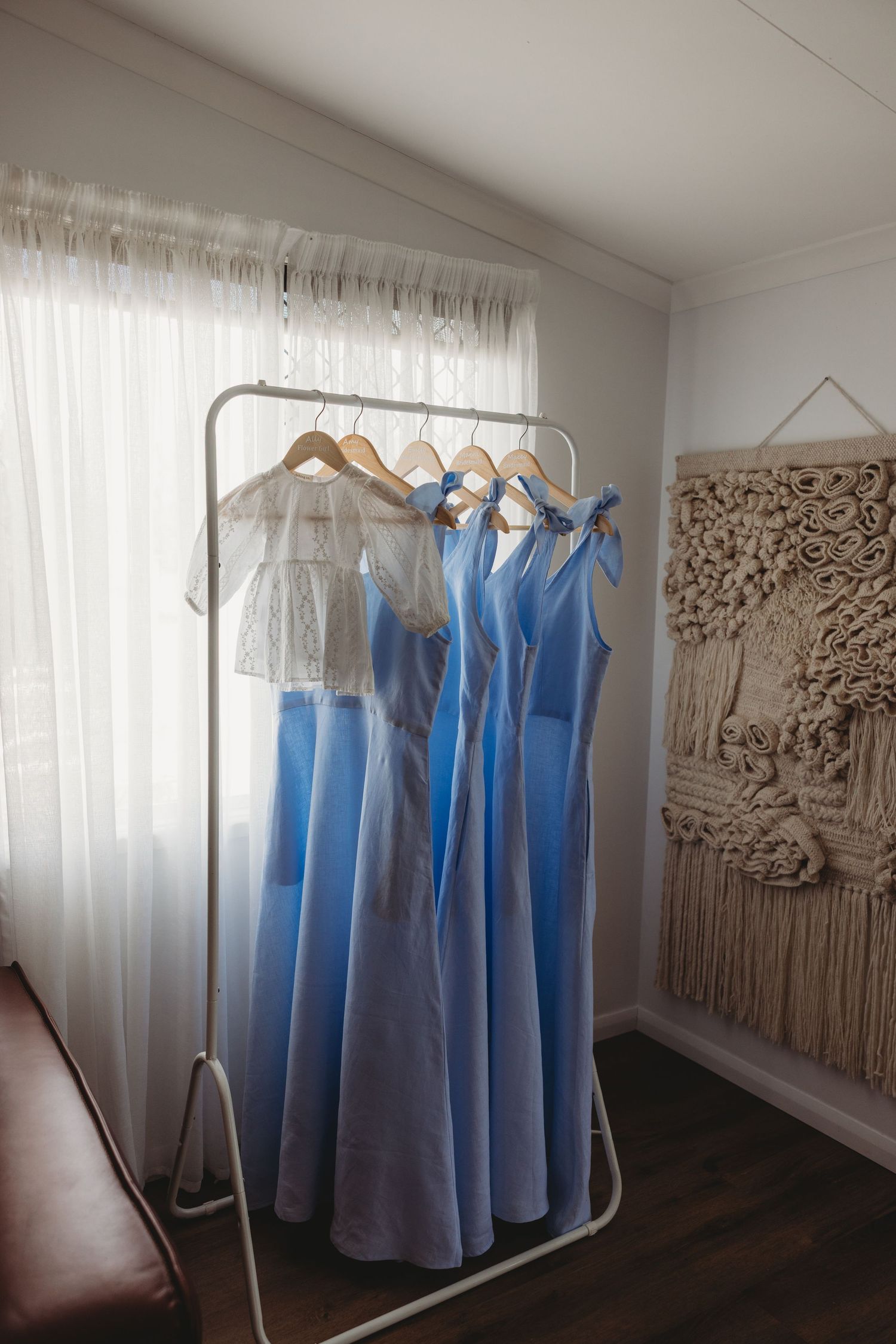 Light blue bridesmaid dresses hanging on a rack against white curtains and macrame wall decor.