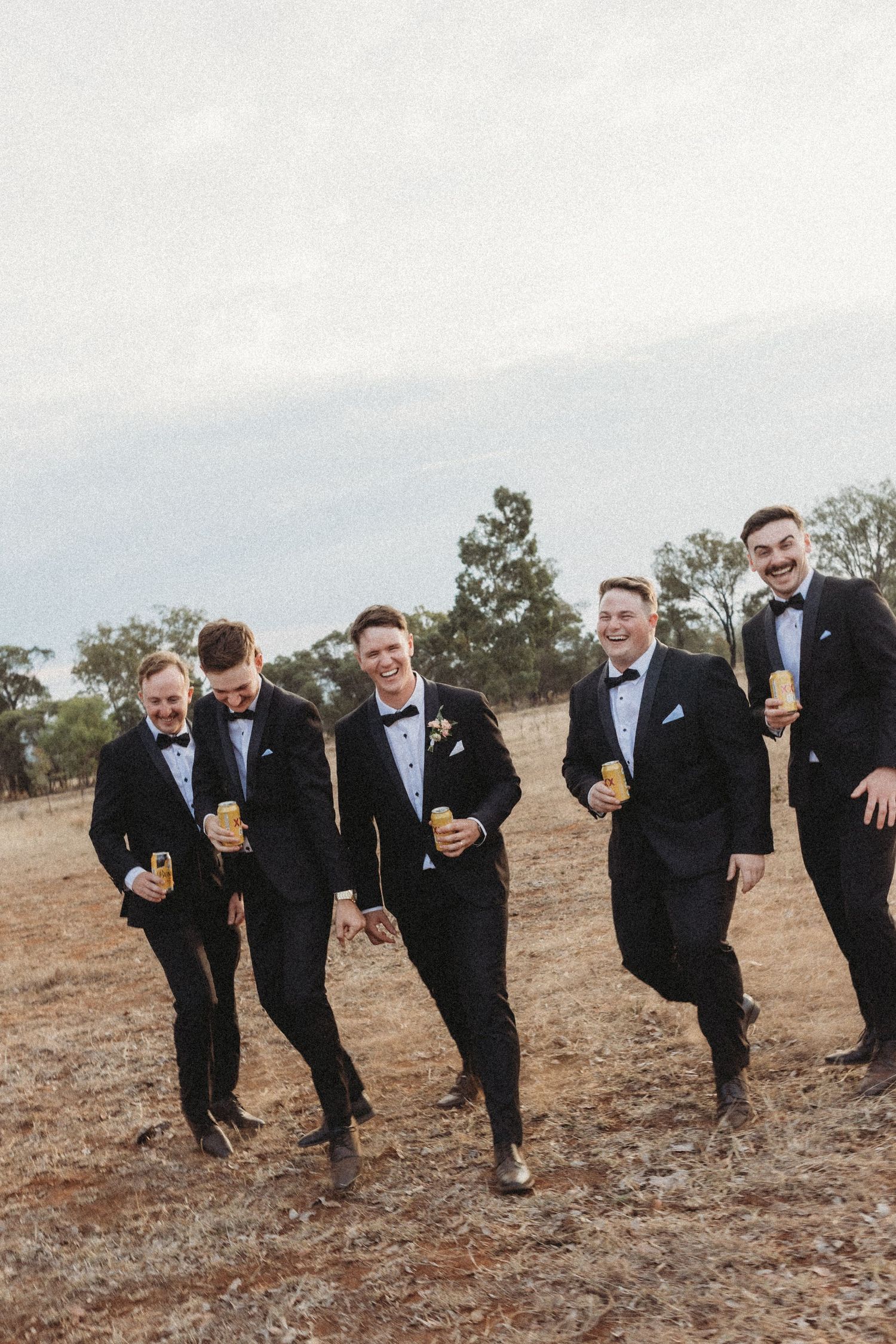 Groomsmen in black tuxedos pose playfully leaning to one side in an outdoor setting.