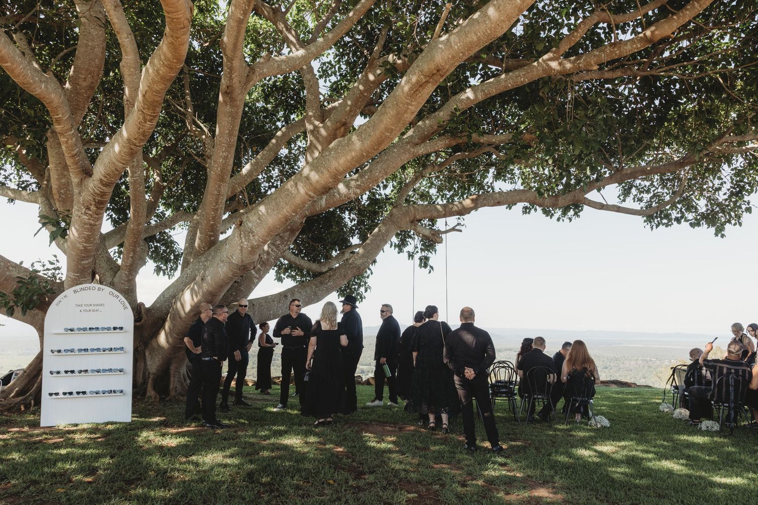 A panoramic view of a gathering under large trees with headstones visible in the background.