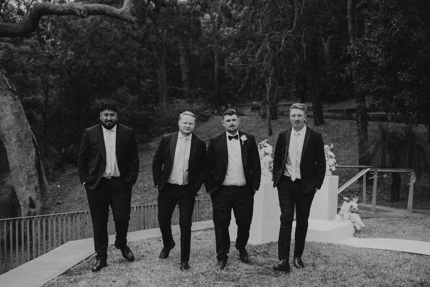 Four groomsmen in black tuxedos and bowties walk together on a path in a black and white photograph.