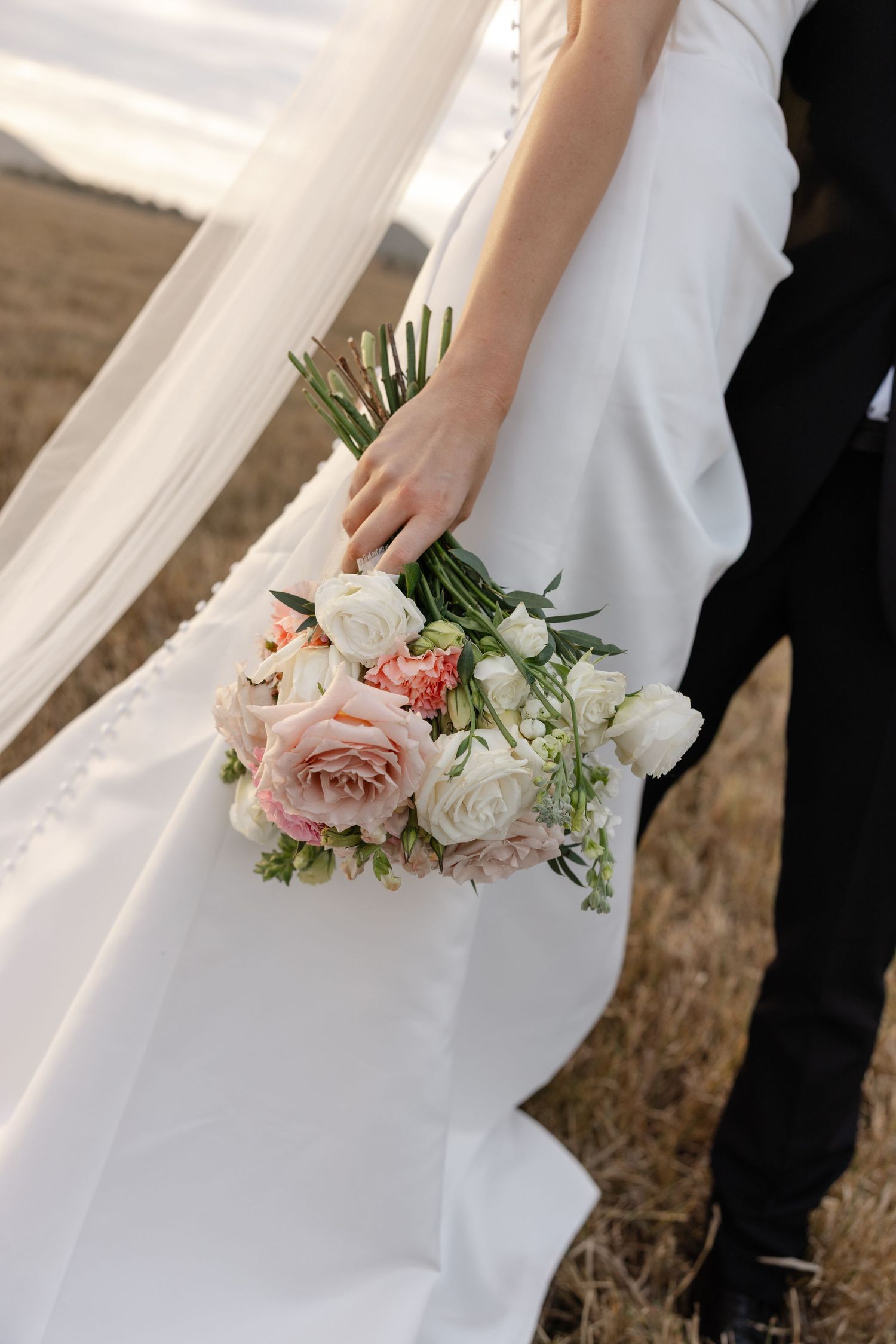 Bride in white gown holding a bouquet of pink and white roses, standing beside groom in black suit outdoors.