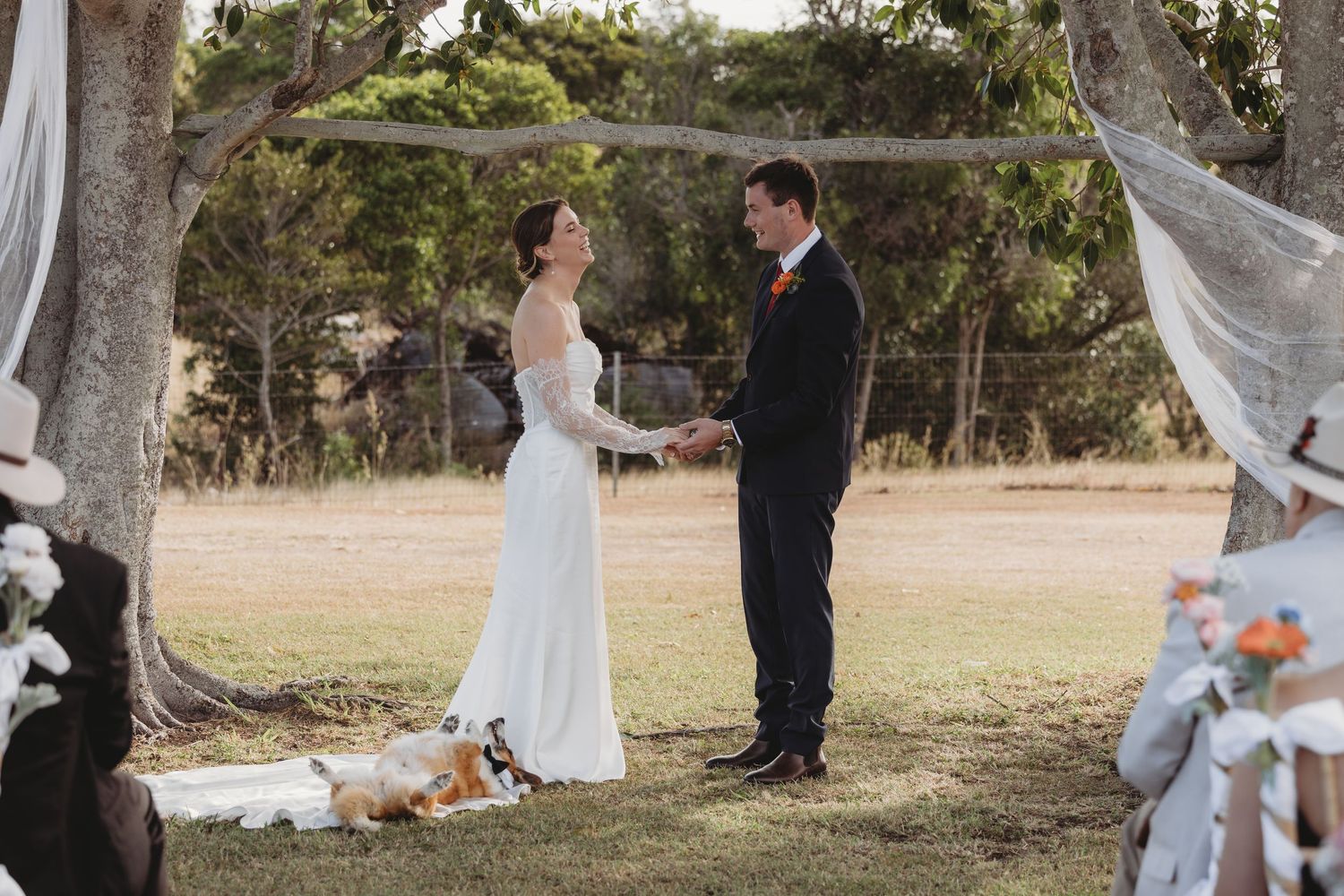 Bride and groom exchange vows at an outdoor wedding altar beneath a rustic tree with a dog resting nearby.