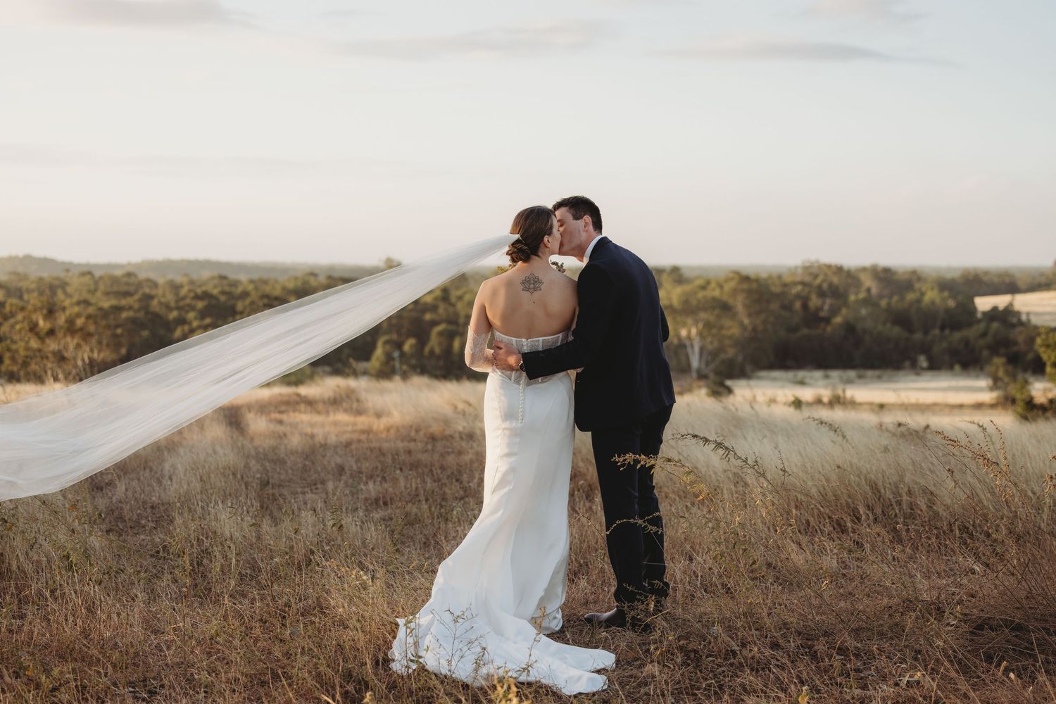 Bride and groom share a romantic kiss in a golden field at sunset, with veil flowing dramatically in the wind.