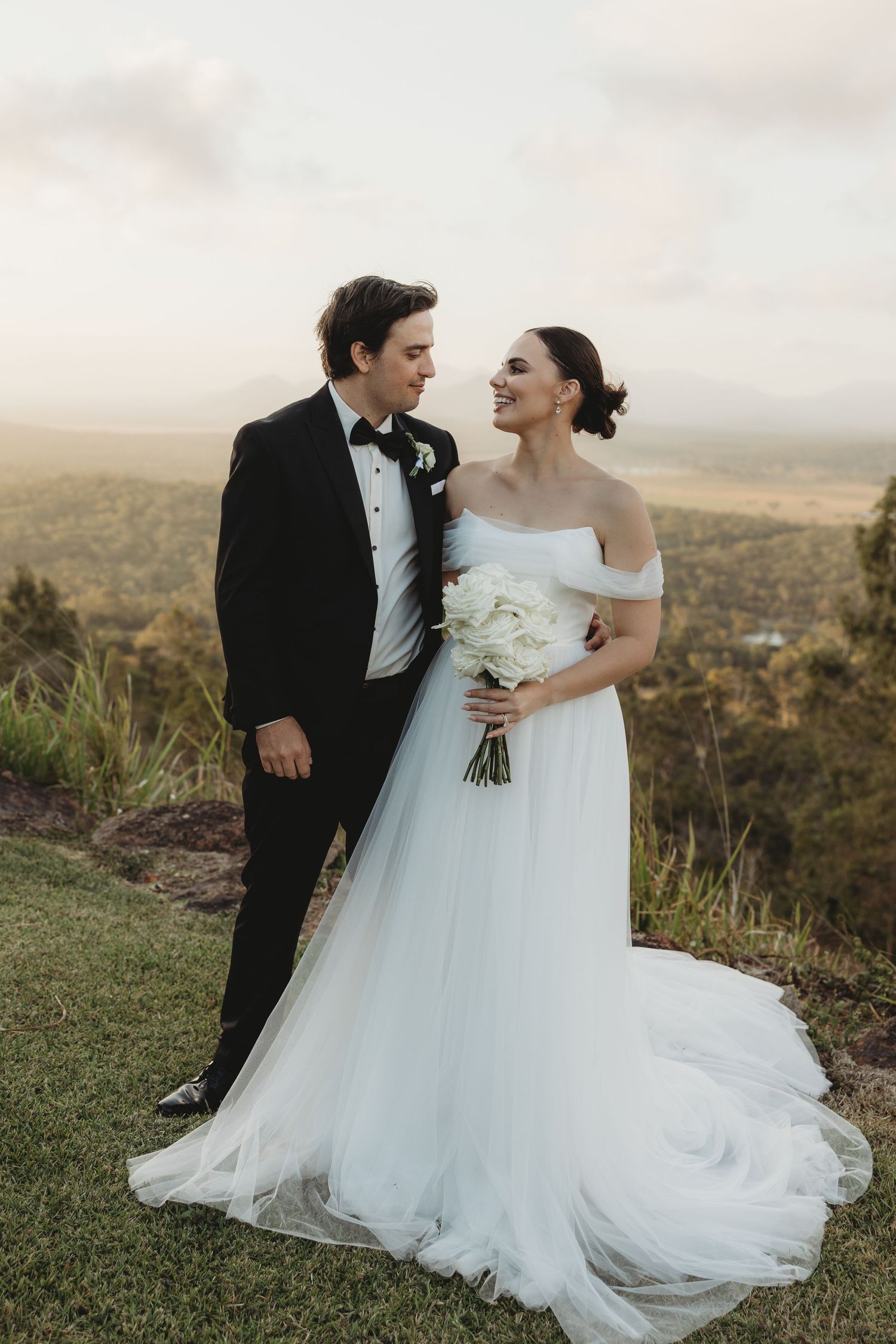 Couple in wedding attire poses on a scenic hilltop with mountain views.