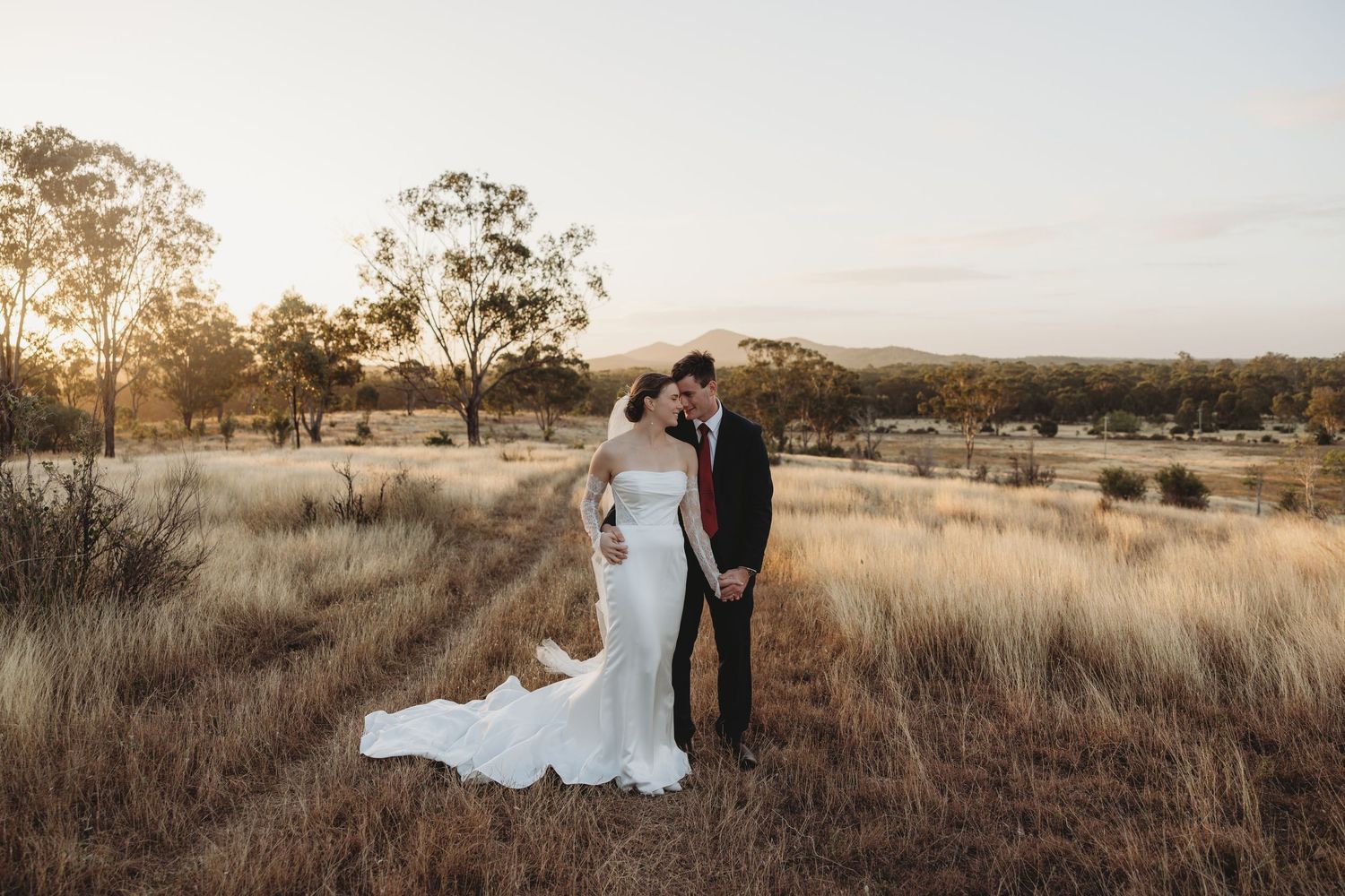 Bride and groom embrace in golden field at sunset with rolling hills backdrop.