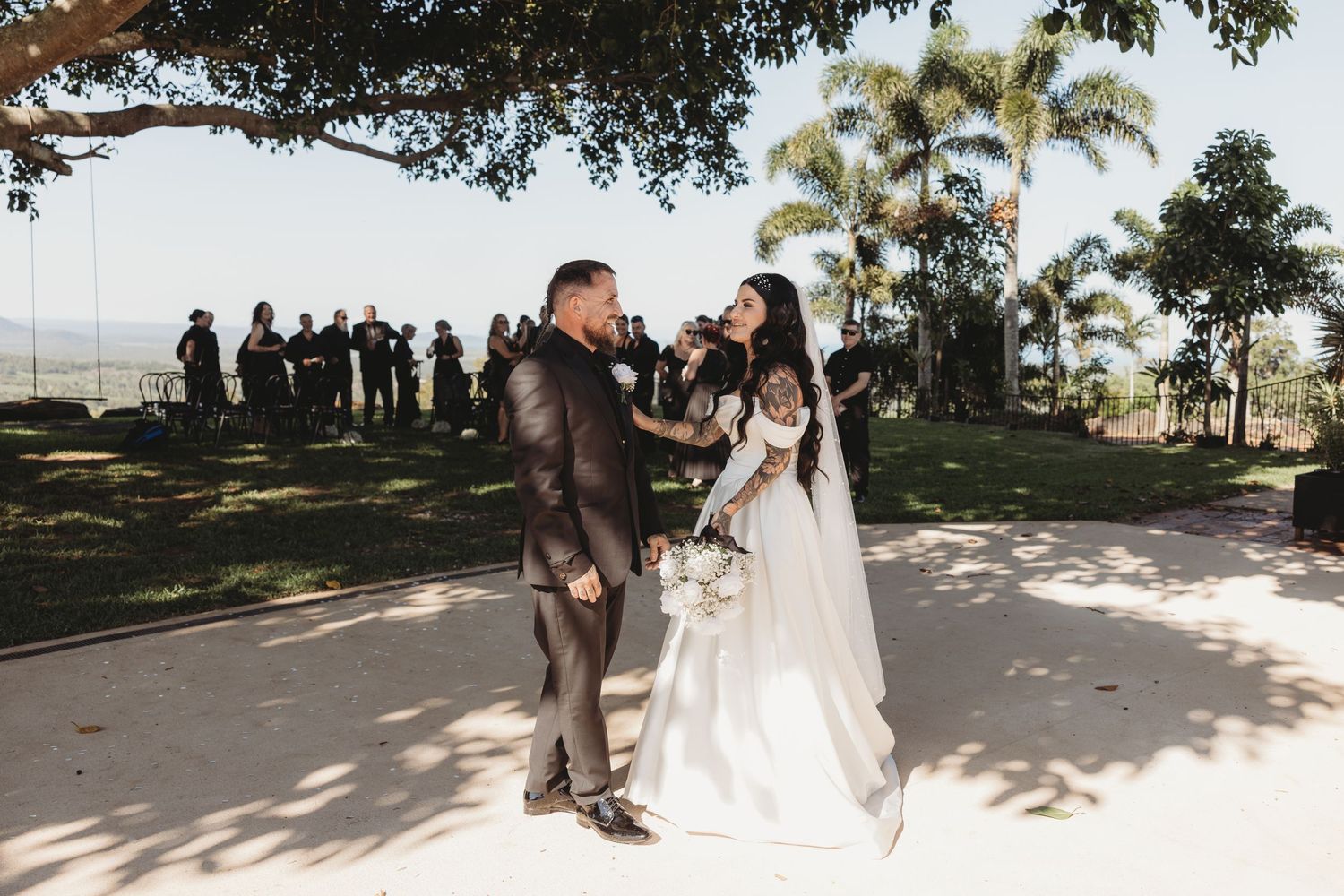 A scenic outdoor wedding ceremony setup with guests beneath towering trees.