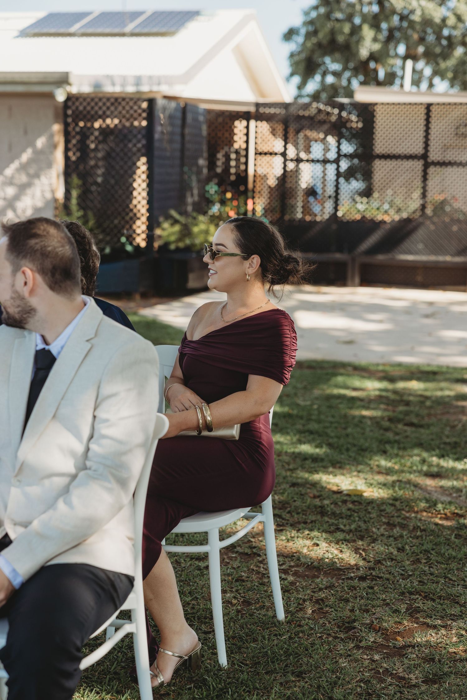 Wedding guests seated in white chairs on a sunny lawn during an outdoor ceremony.