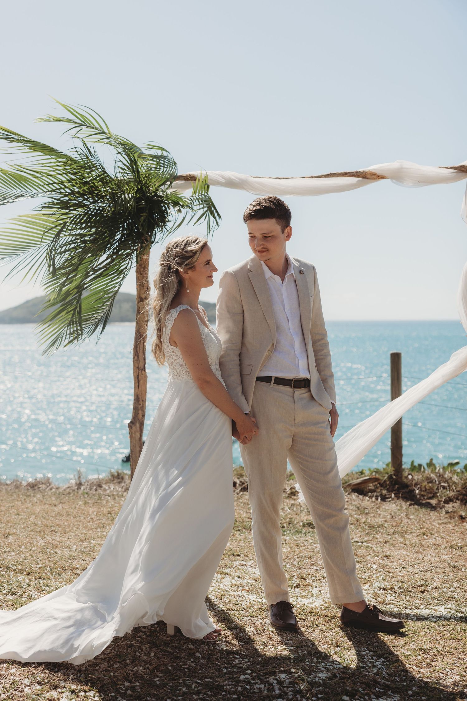 Newlyweds posing for romantic photos by palm trees with ocean backdrop.