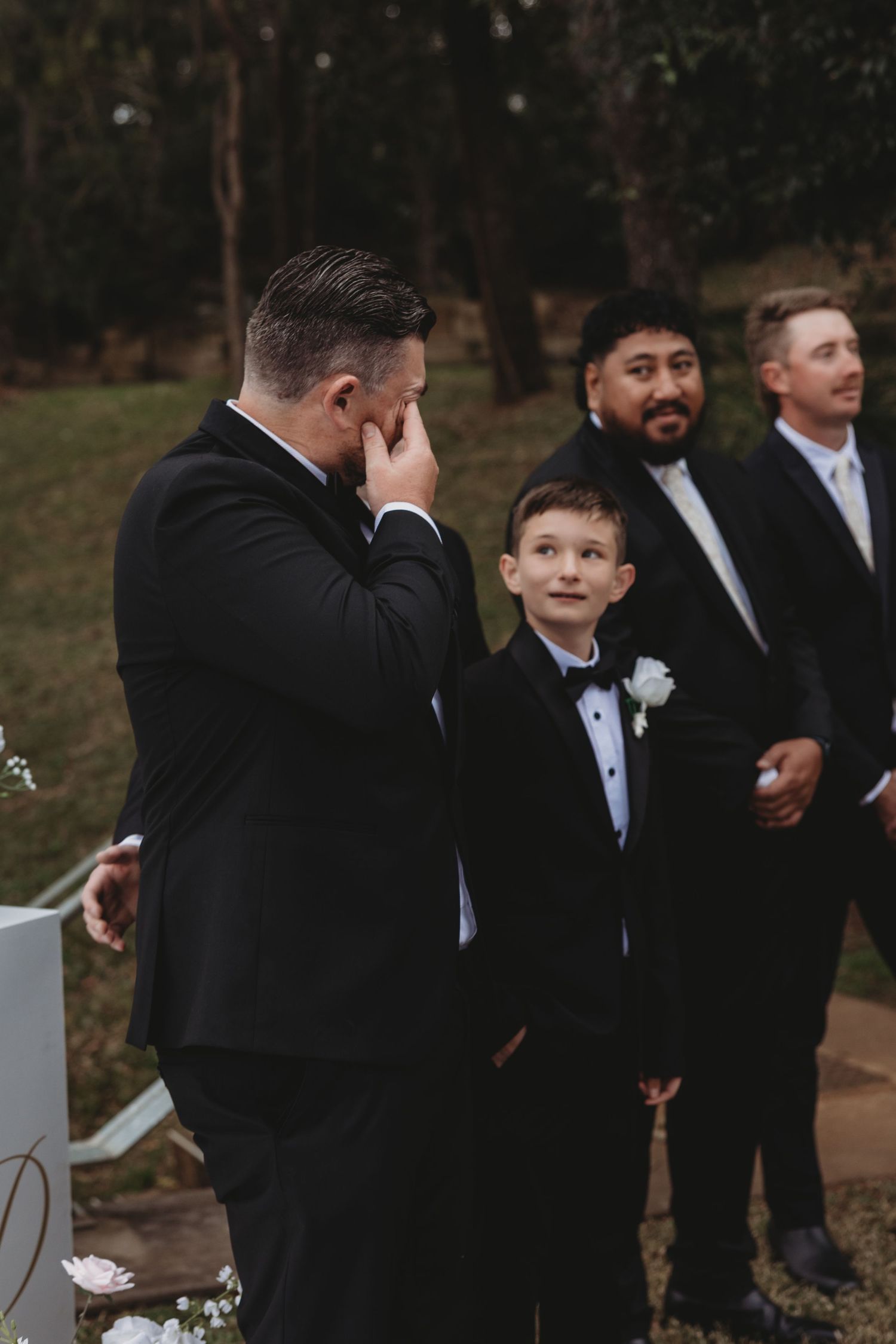 Groomsmen in black tuxedos share candid moments and laughter during outdoor wedding photo session.