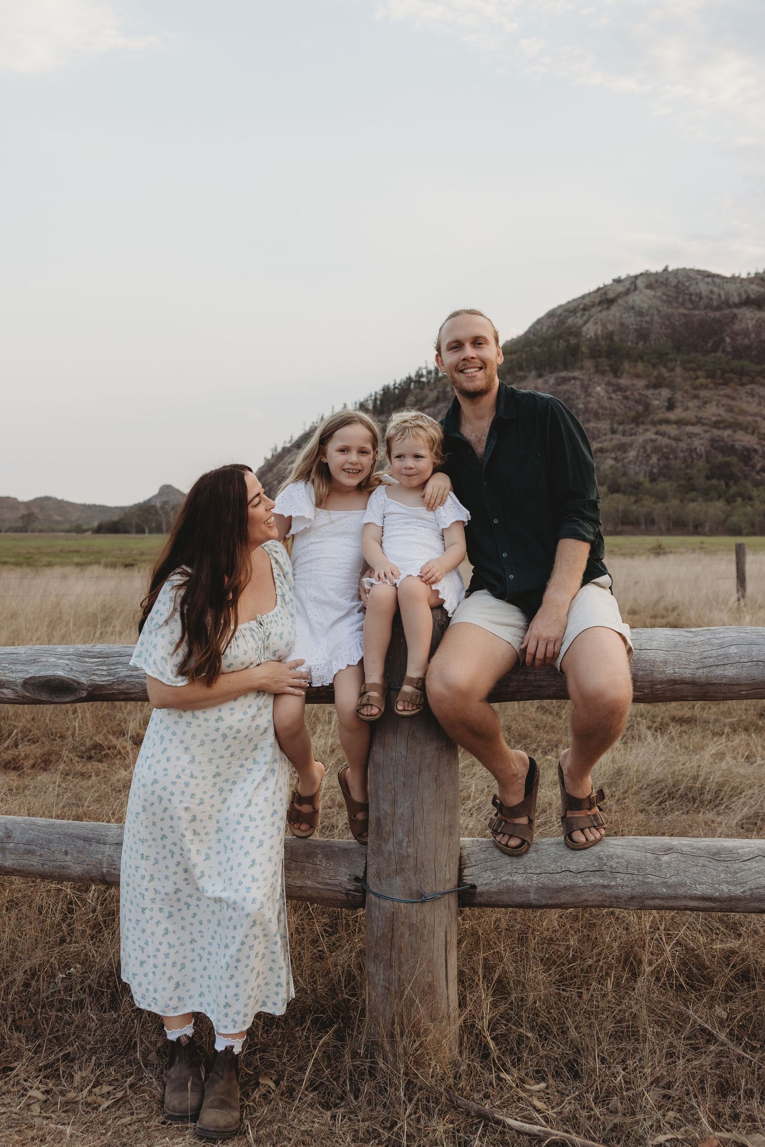 Family sitting on wooden fence with mountain landscape in background.