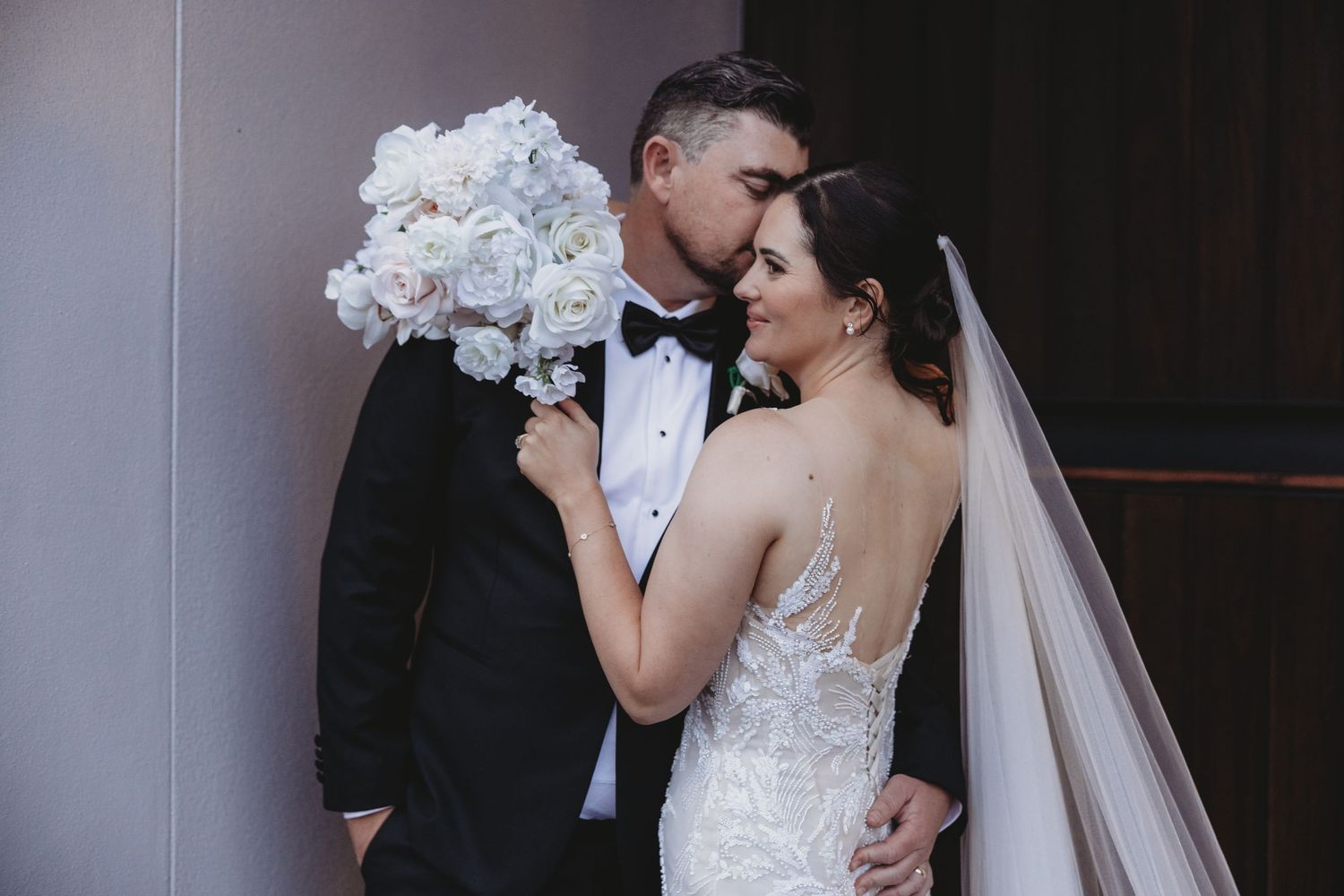 Elegant wedding couple shares intimate moments in a dark indoor setting with white floral bouquet.