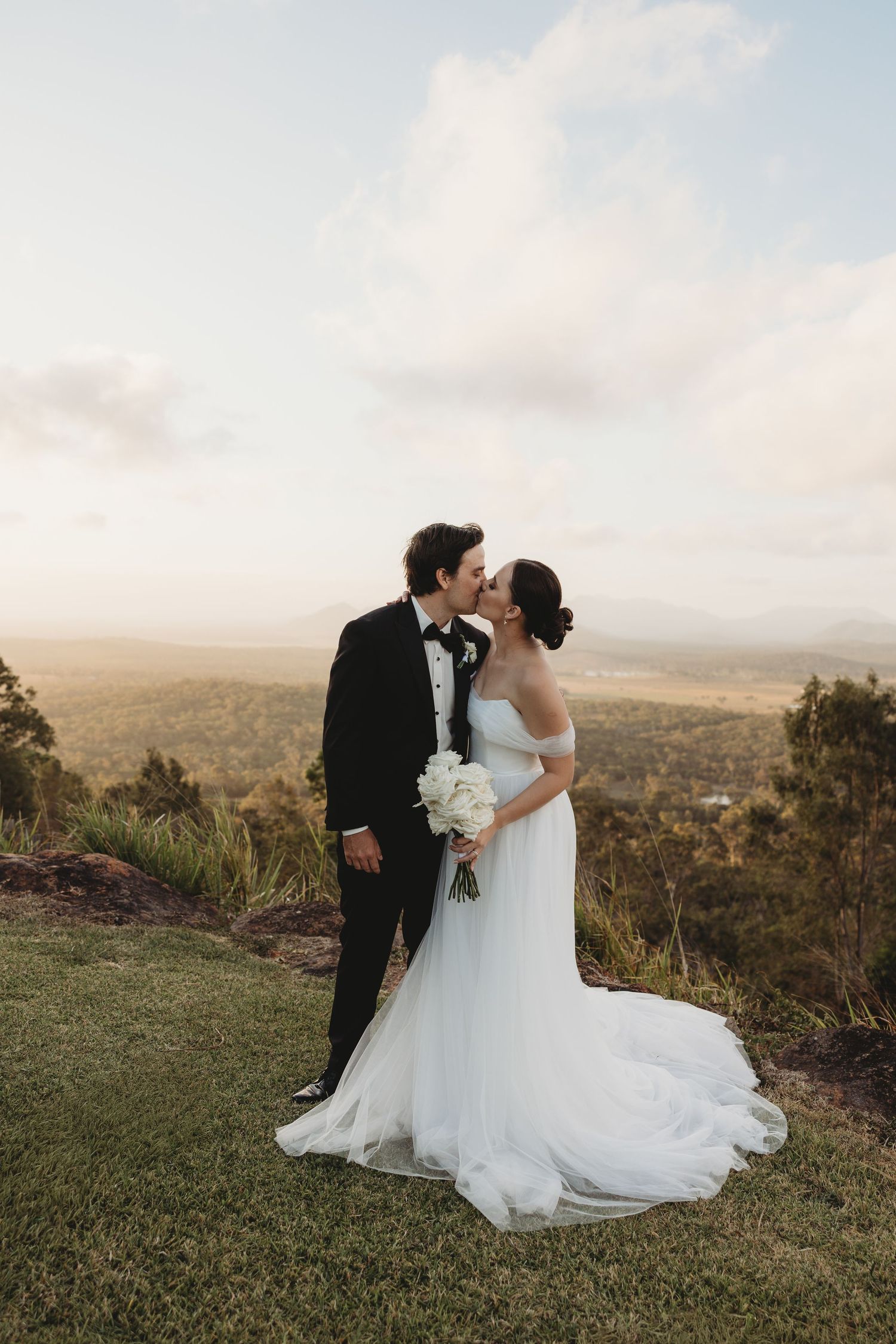 Newlyweds share romantic moment on grassy hilltop with panoramic valley views.