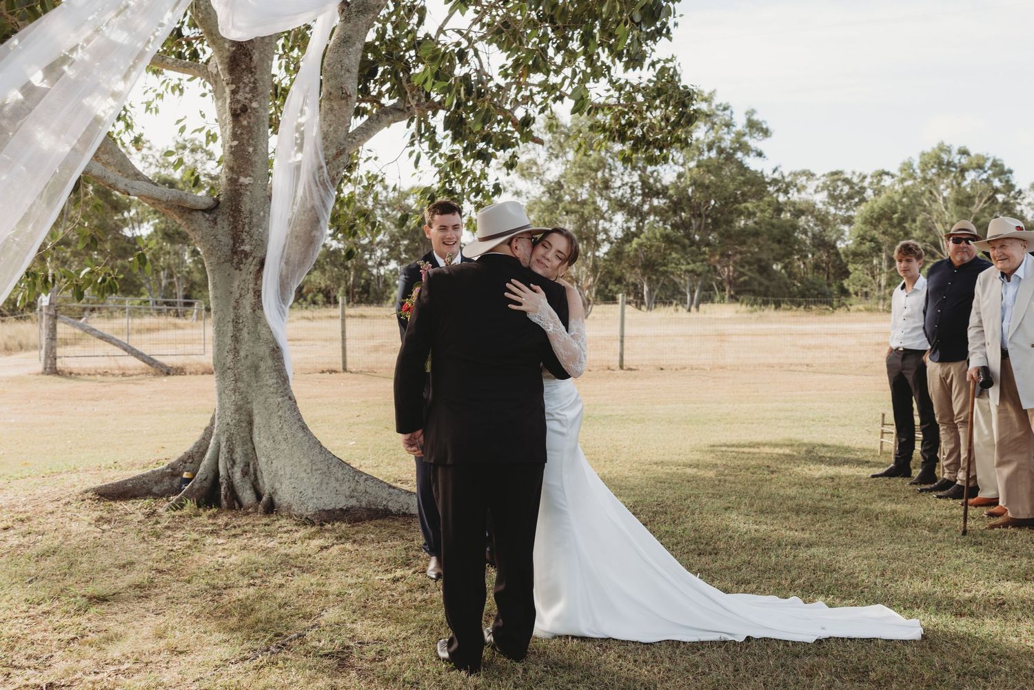 Bride hugging guest during outdoor farm wedding ceremony beneath decorated tree.