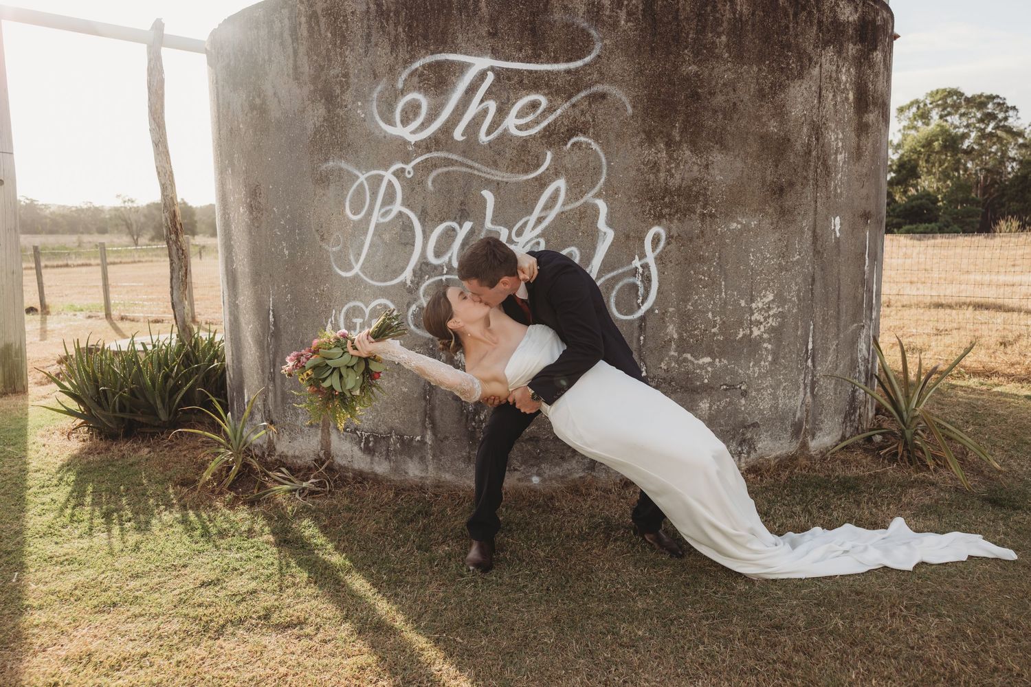 Bride and groom share a romantic dip kiss in front of a rustic silo with The Barbs written in white calligraphy.