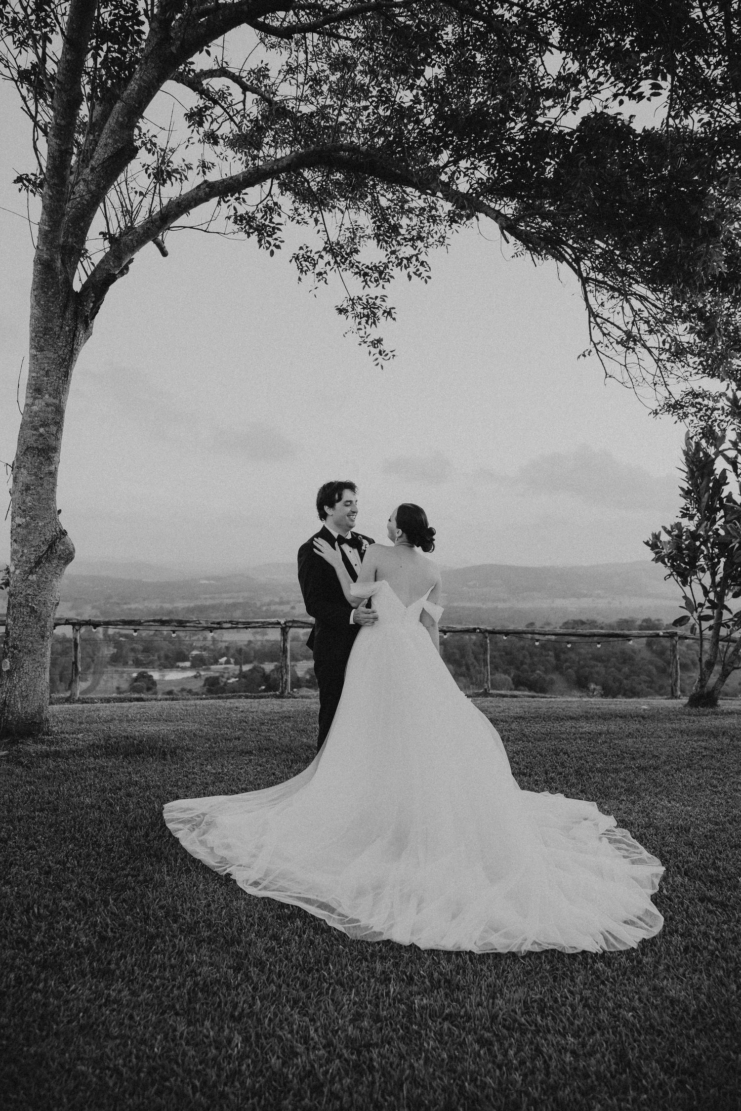 Black and white wedding portrait under a large tree with a dramatic flowing dress and mountain backdrop.