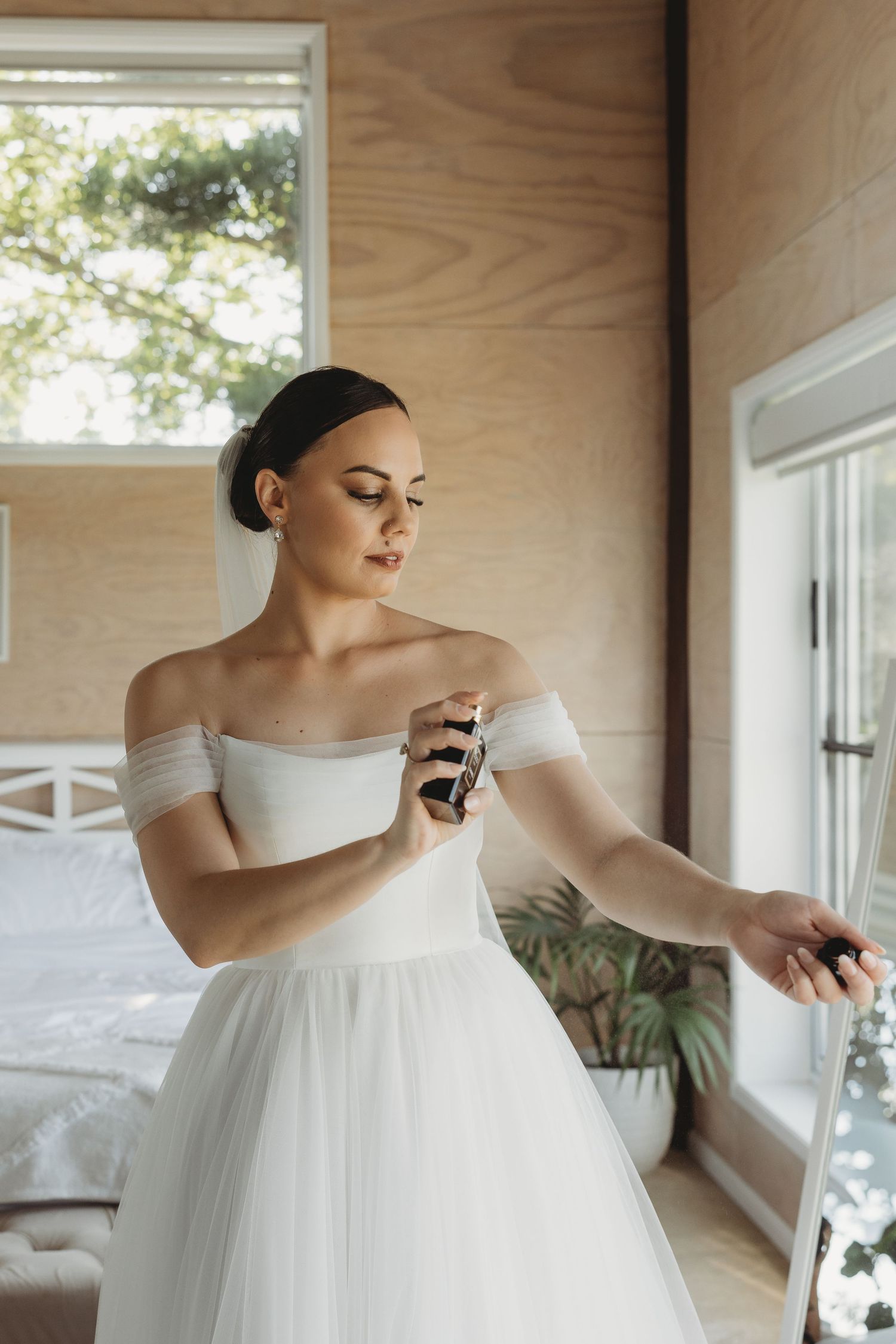 Bride in off-shoulder ballgown preparing in a sunlit room with wooden walls.