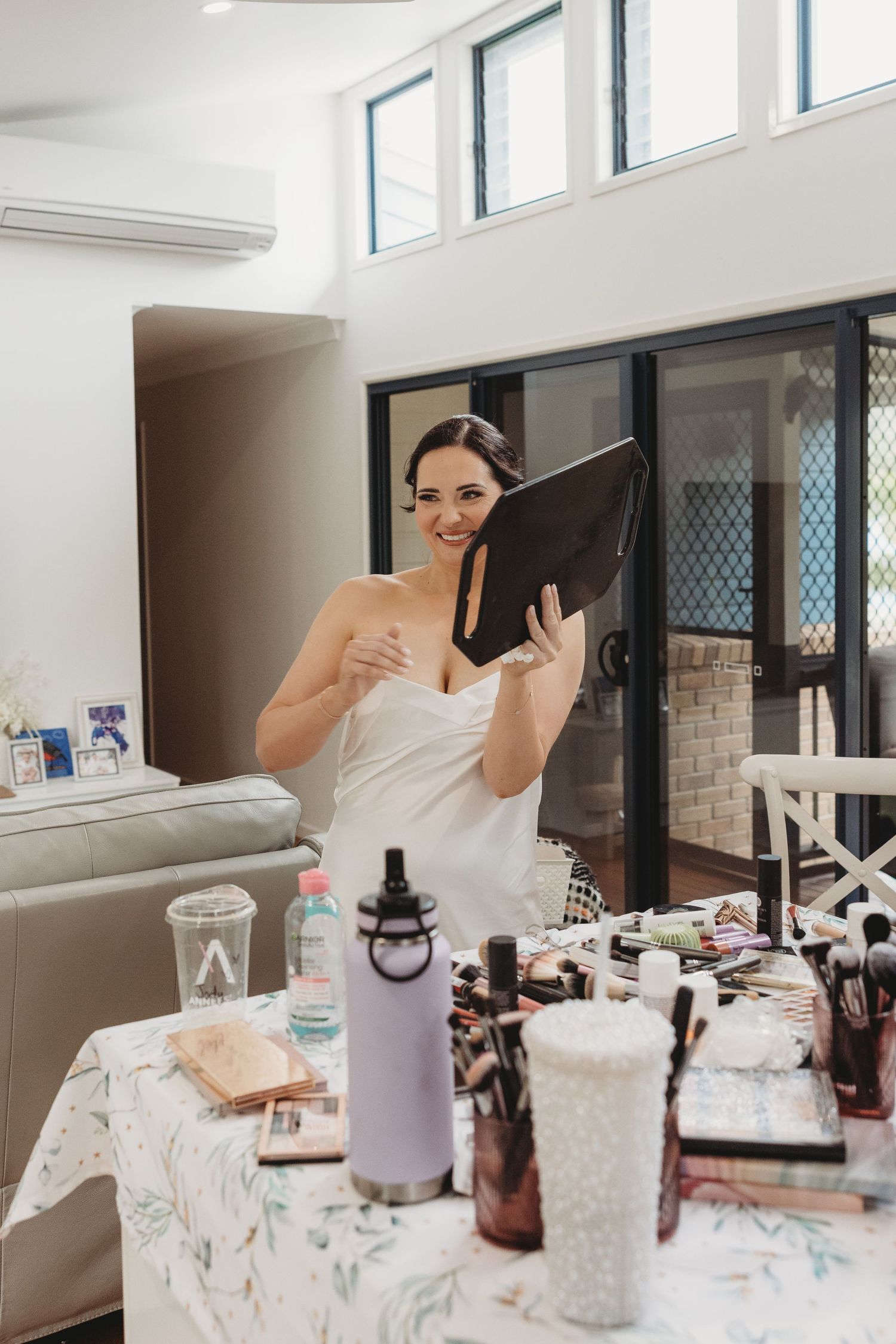 Someone in a white strapless dress stands at a table with makeup and beauty products during preparation.