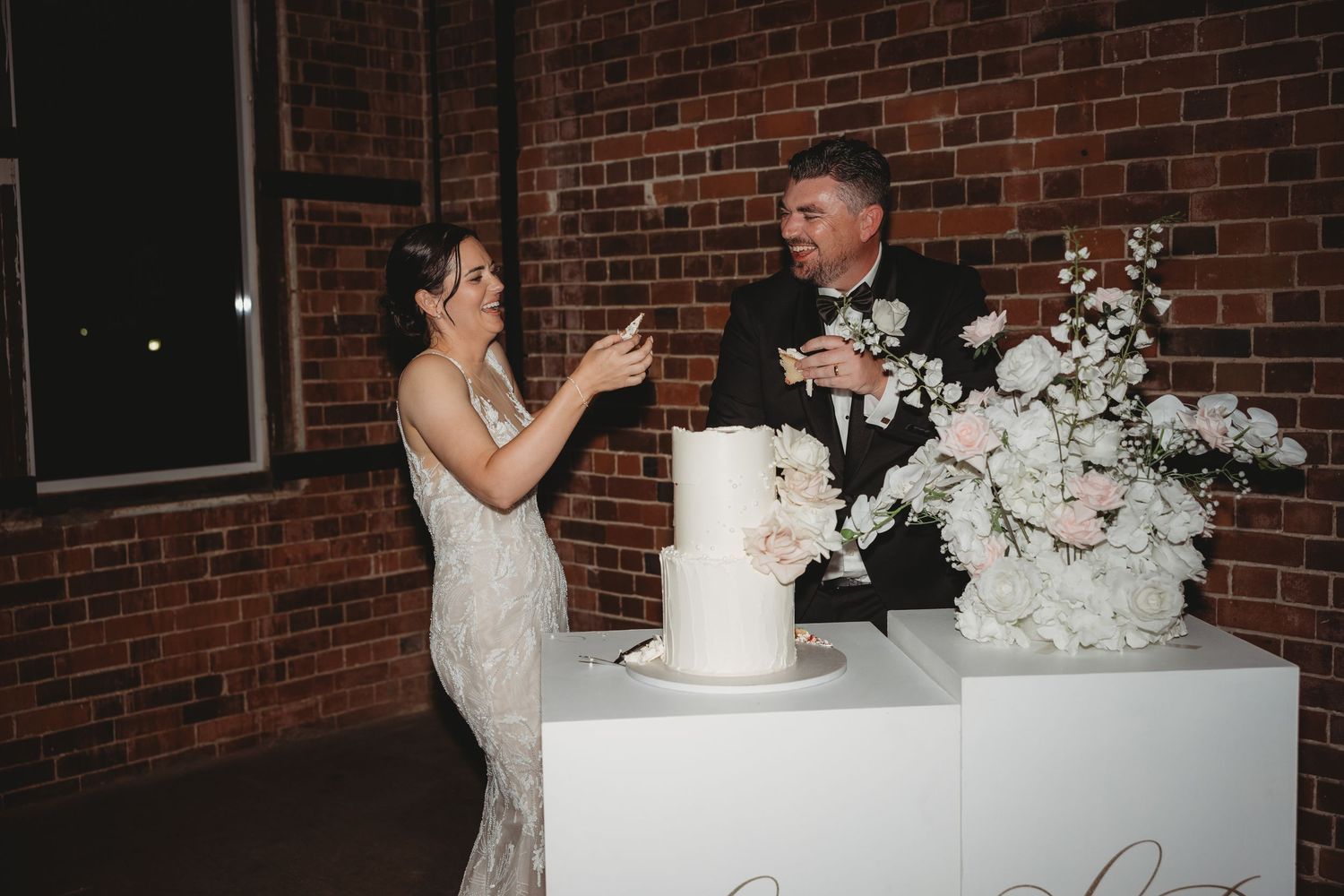 Couple sharing a moment near their elegant white wedding cake and floral display against a brick wall.