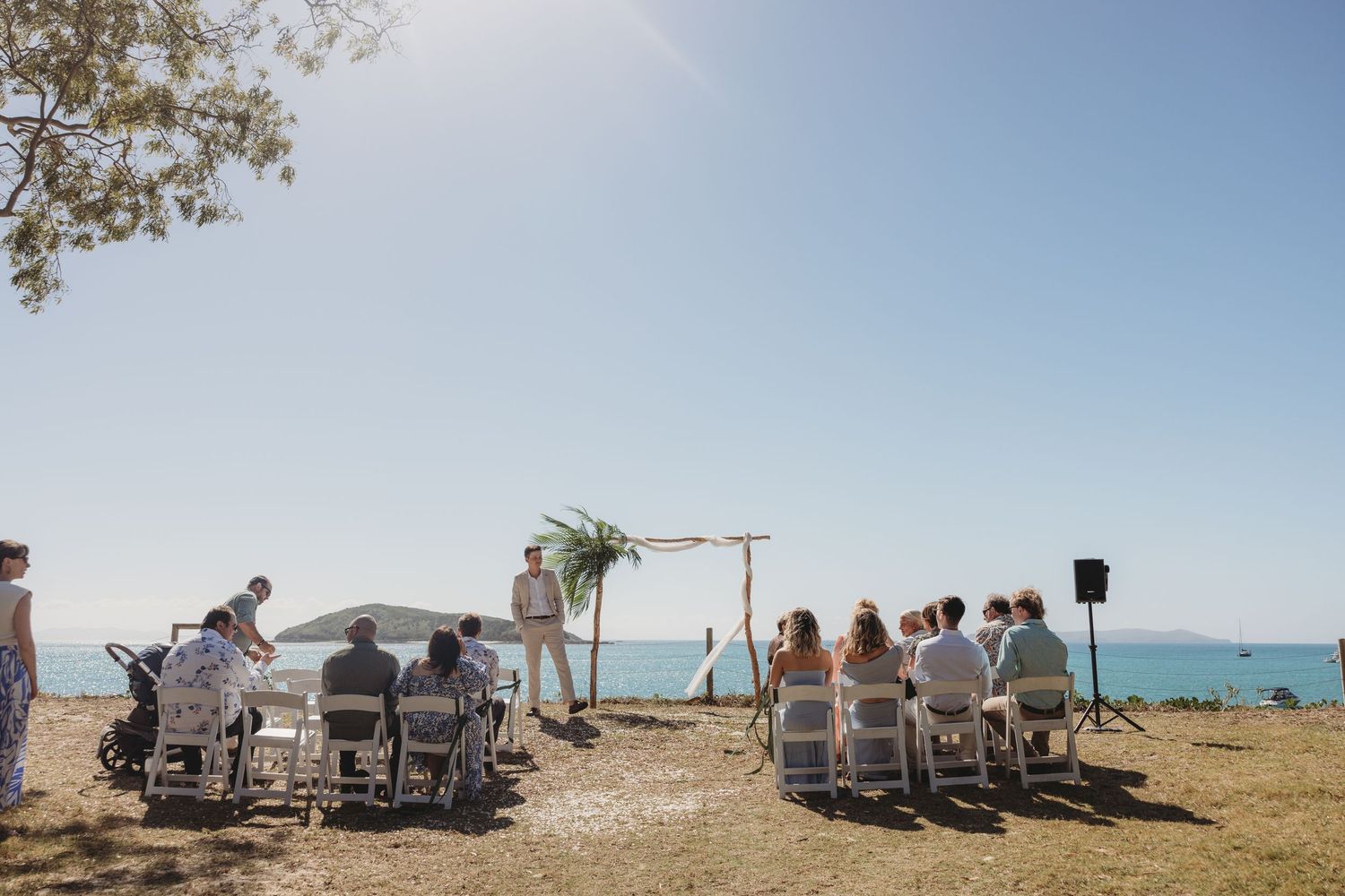 Panoramic view of guests seated at a beachside wedding ceremony with white chairs and ocean backdrop.
