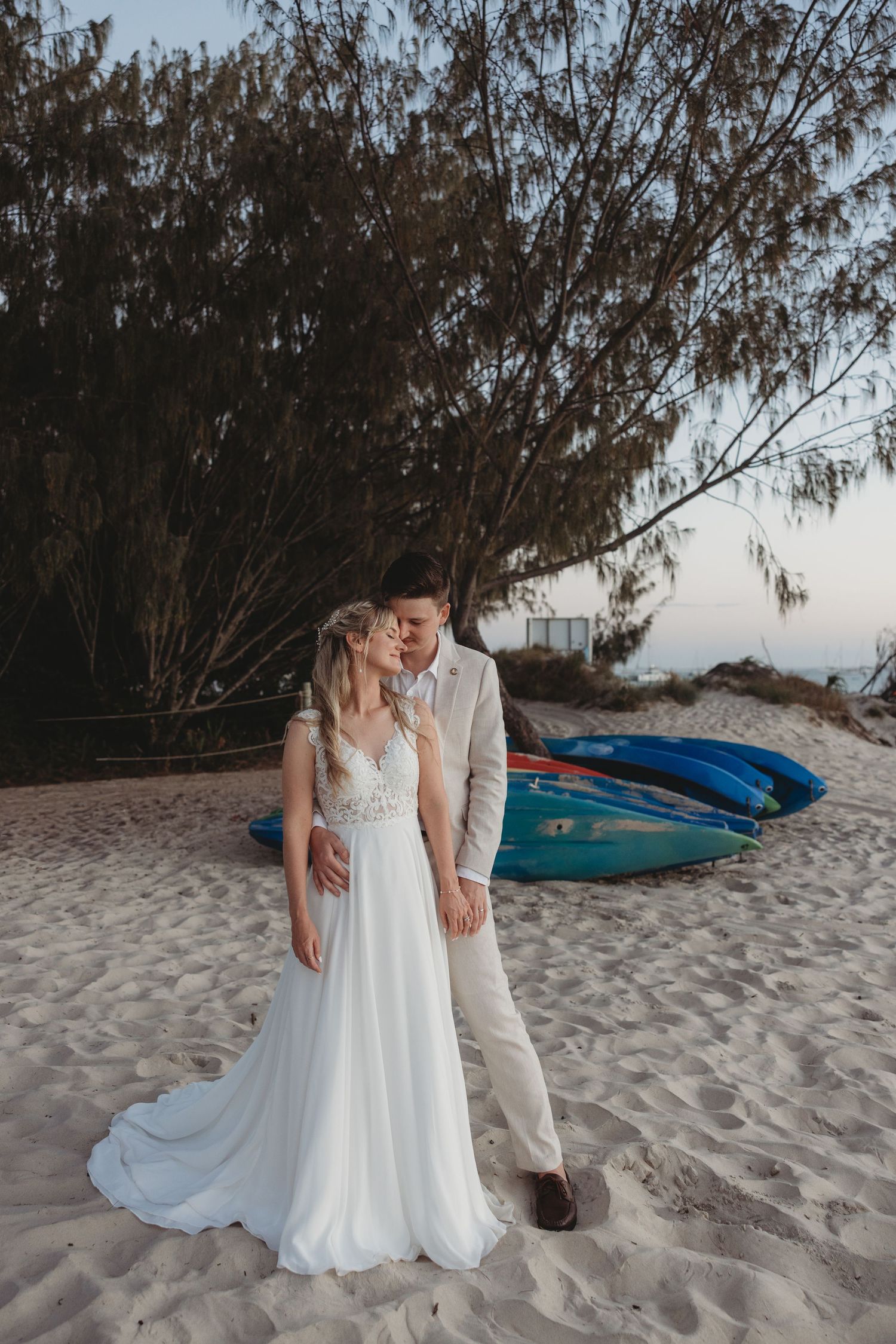 Newlywed couple poses on sandy beach with kayaks in background at sunset.