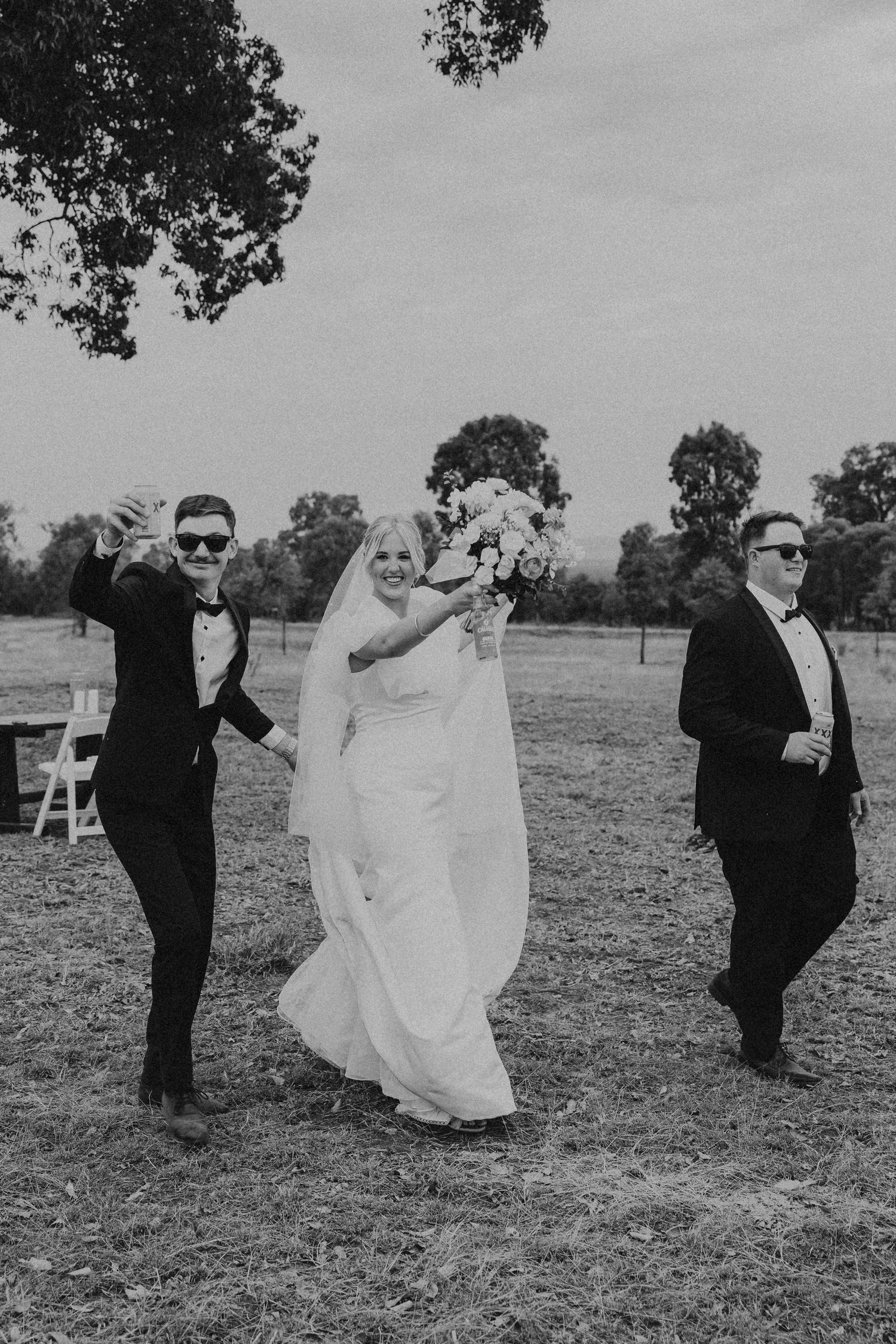 A wedding party poses playfully in a rustic field wearing formal attire in black and white.