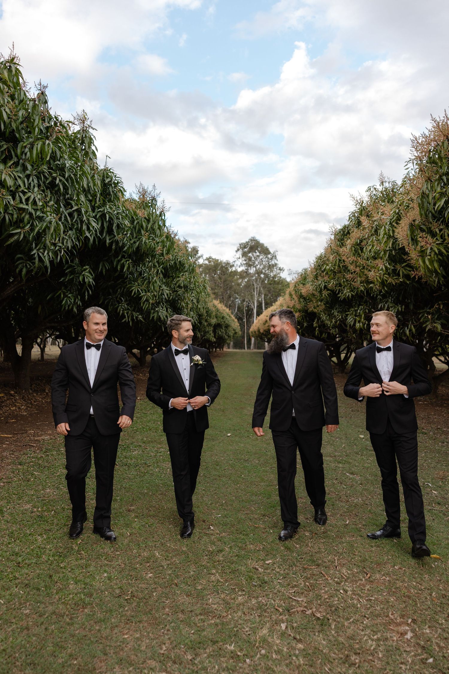 Four groomsmen in black tuxedos with bow ties walk together outdoors on green grass between rows of trees.