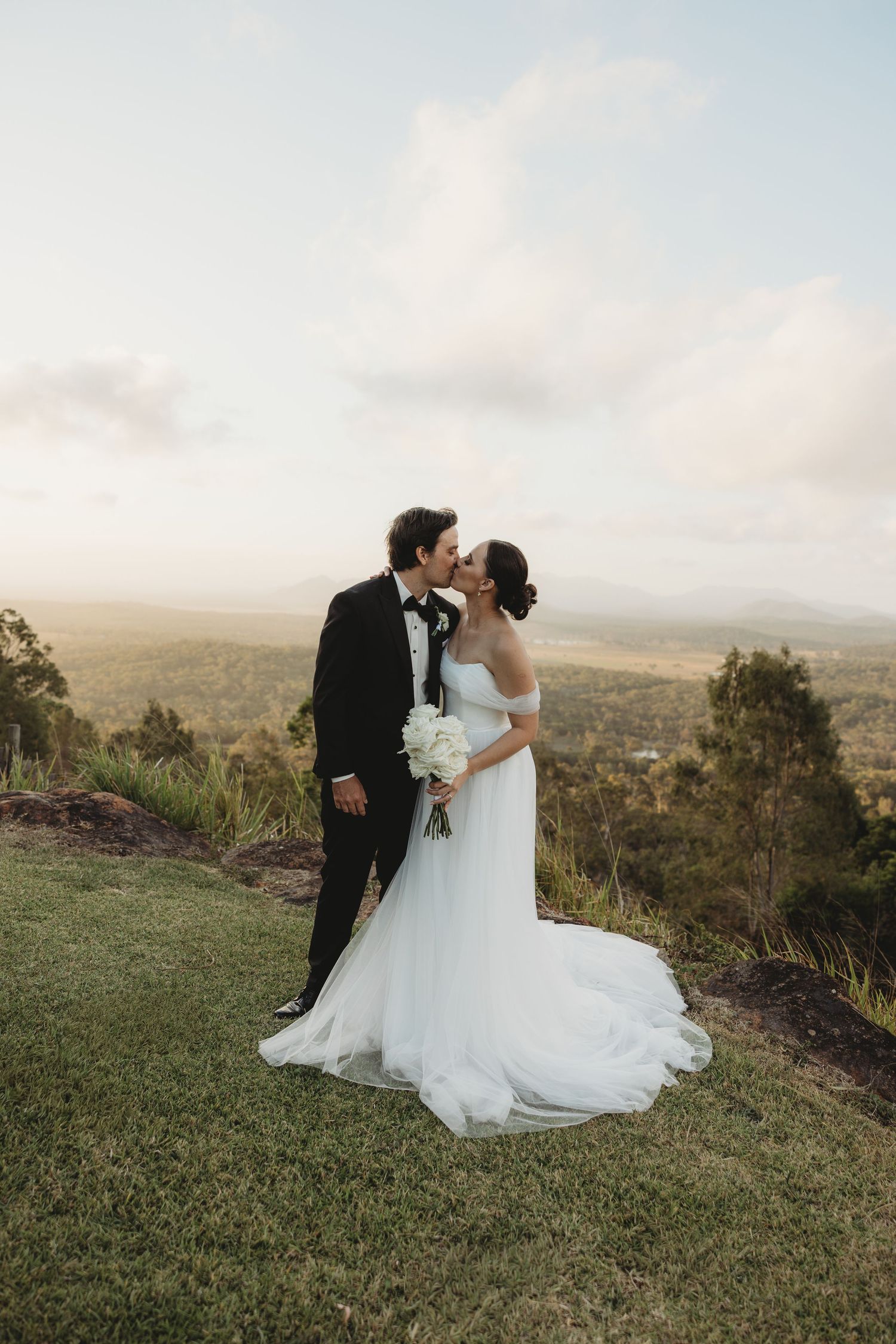 Newlyweds share an intimate moment on a grassy hilltop with scenic mountain backdrop.