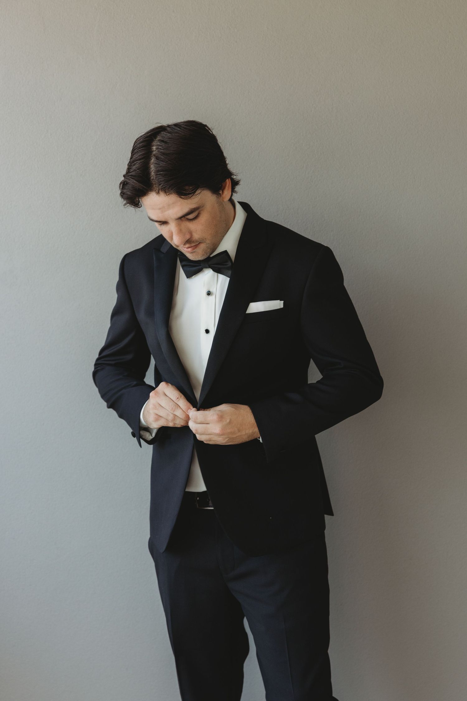 A person in a black tuxedo adjusts their bowtie while getting ready for a formal event.