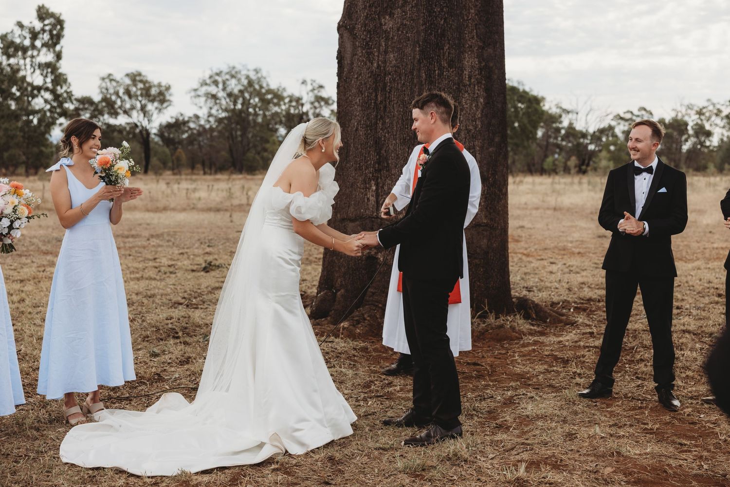 Wedding ceremony participants gathered around a large tree in a rural setting.