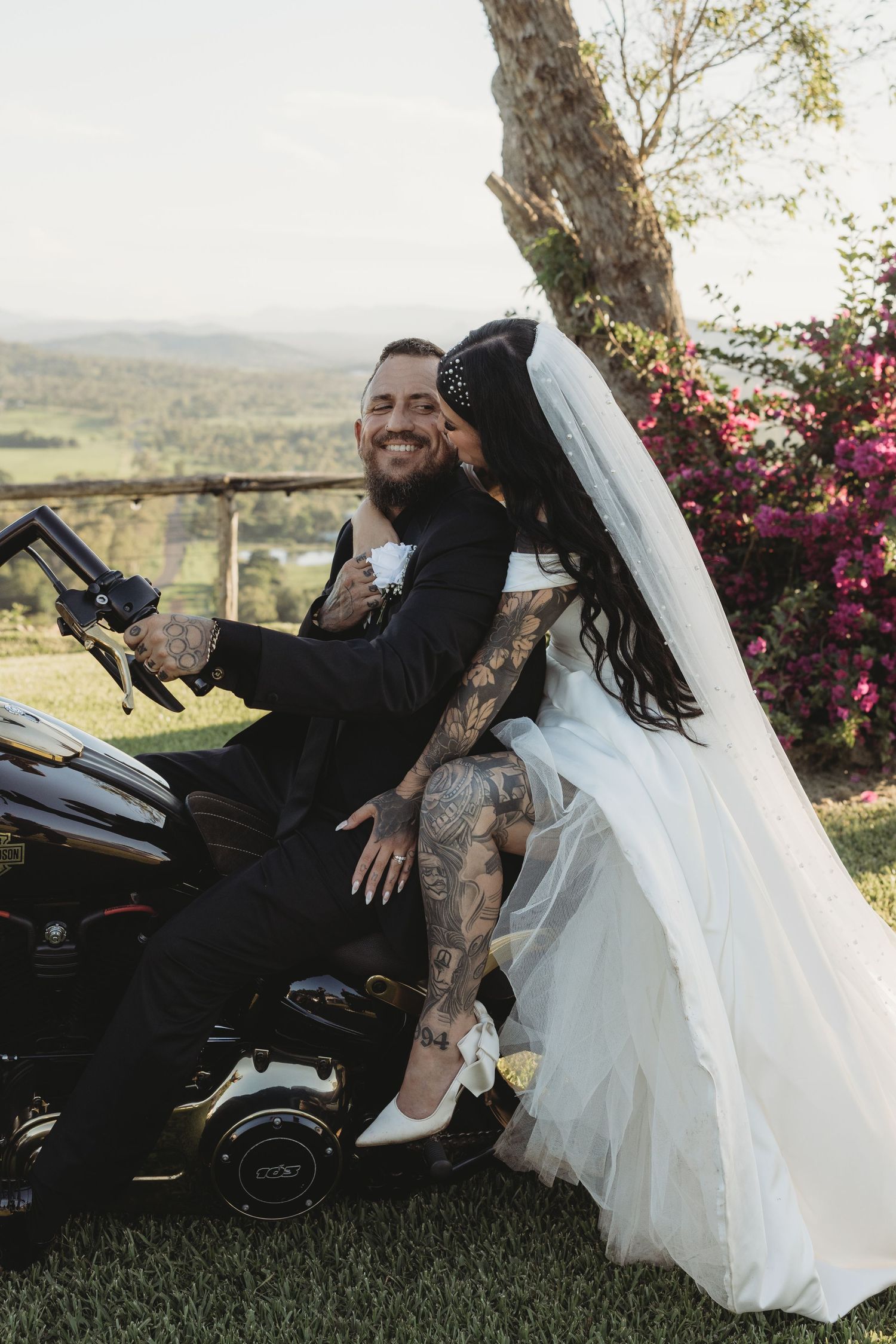 A newlywed couple poses on a motorcycle with a scenic overlook and flowering trees in the background.