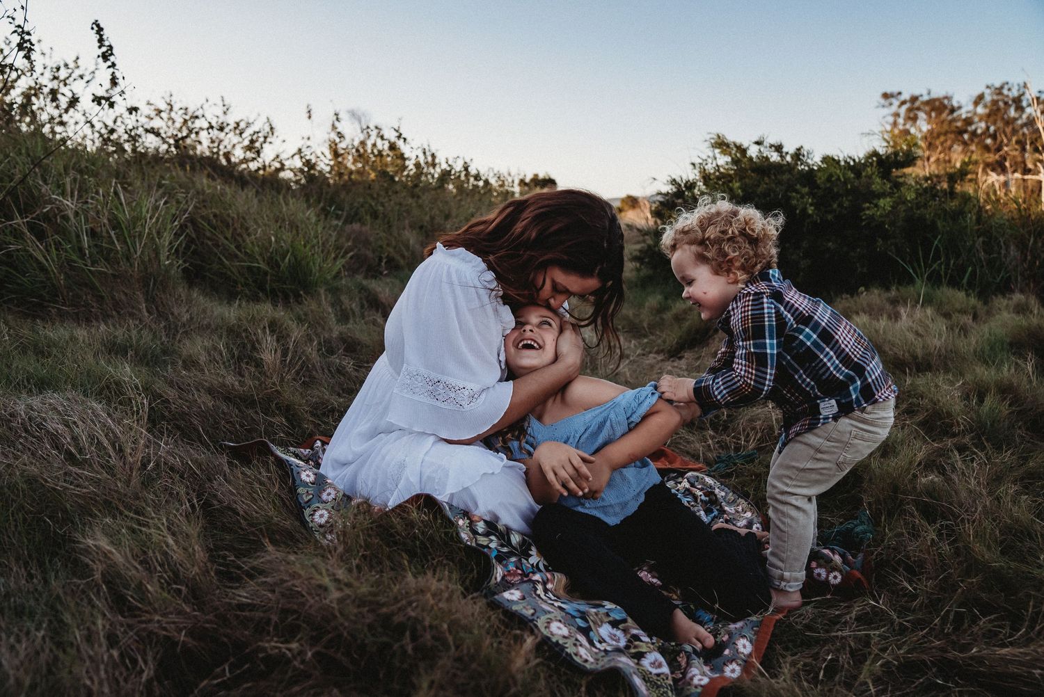 Family enjoying playful moment together on blanket in grassy field at sunset.