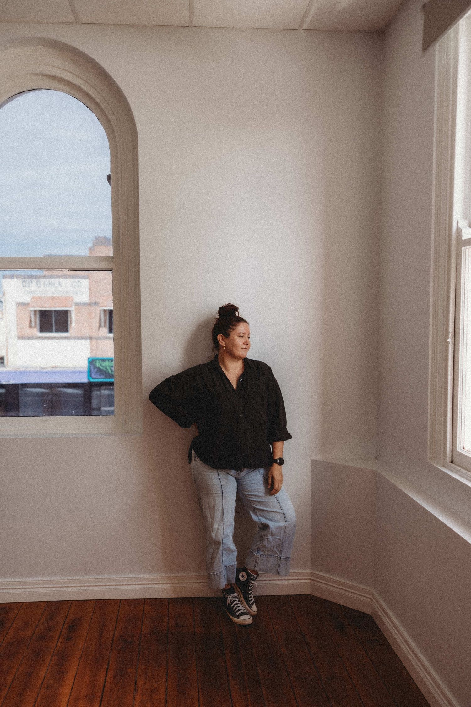 Woman in black top and light jeans leaning against white wall near arched window in bright minimalist room.