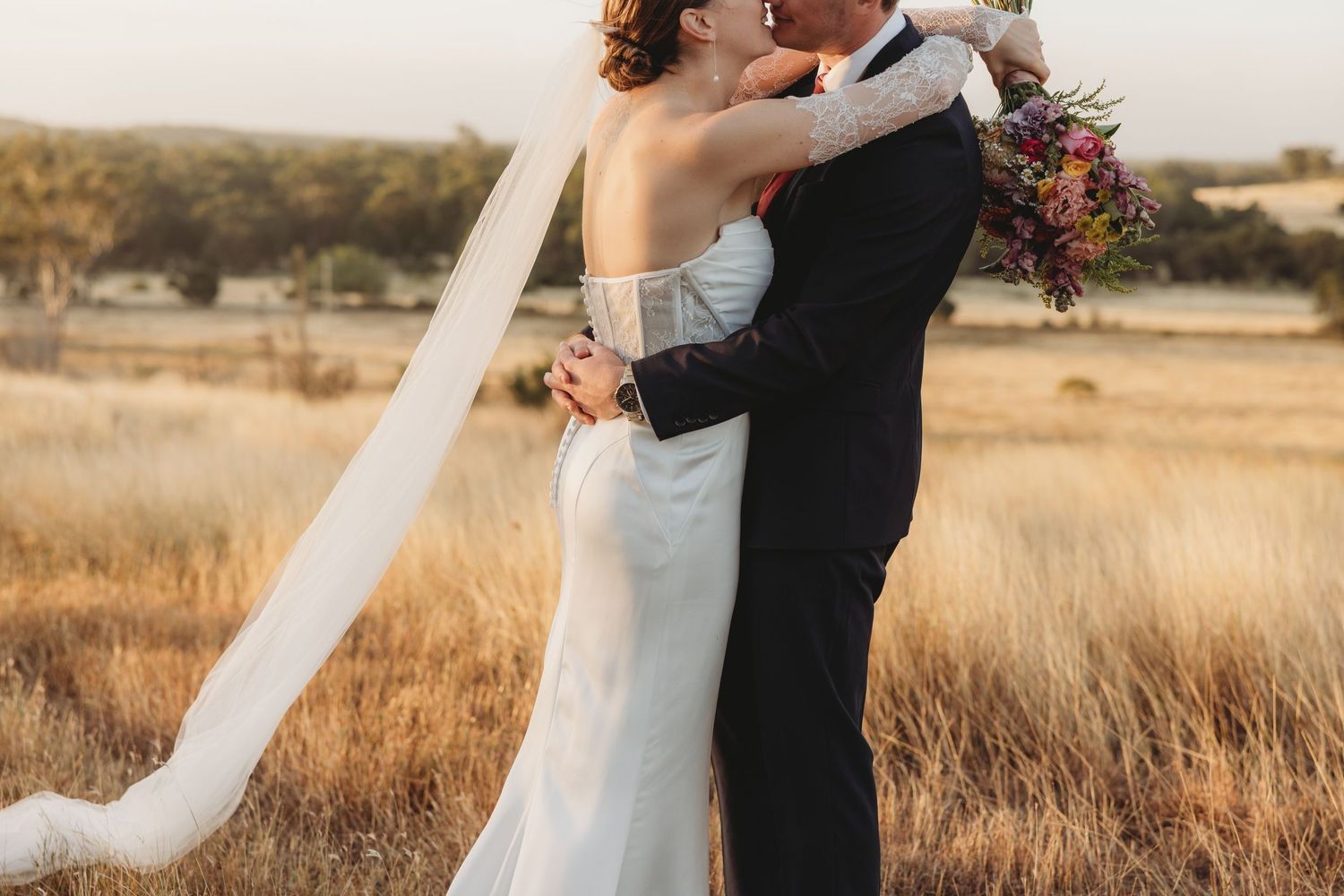 Bride and groom embrace and kiss in a golden field at sunset, bride holding a colorful bouquet with veil flowing.