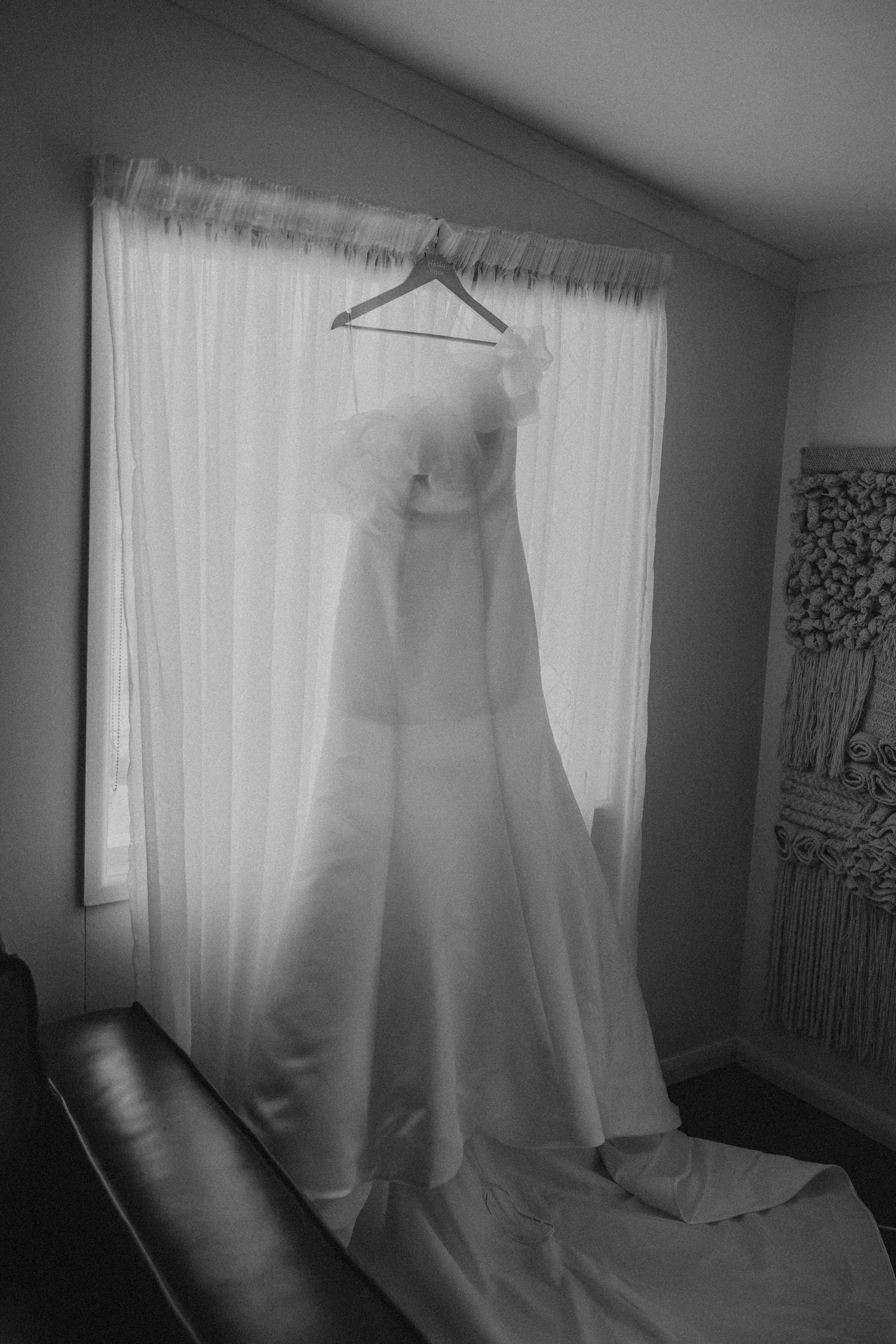 Wedding dress hanging in front of window with sheer white curtains in moody black and white.