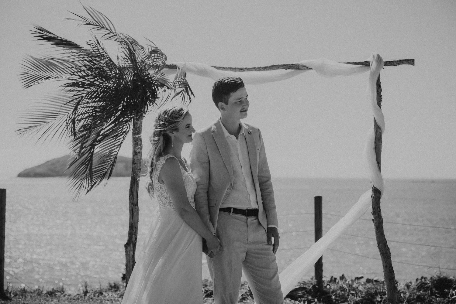 Black and white photo of couple posing near palm trees by the ocean.