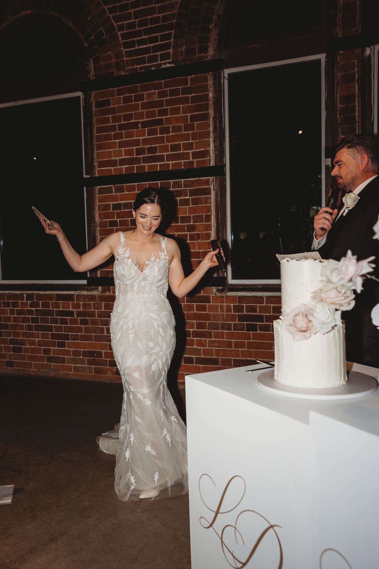 A bride and groom cut their elegant white wedding cake together at a rustic brick-walled reception venue.