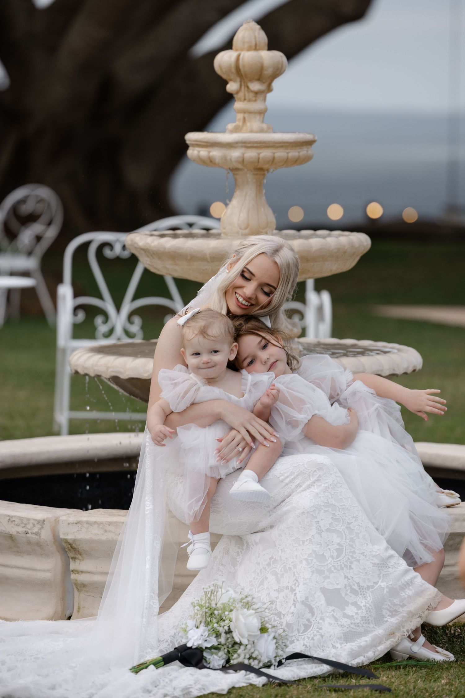 Bride in white wedding gown sits by stone fountain with two young girls in white dresses, sharing a tender embrace outdoors.