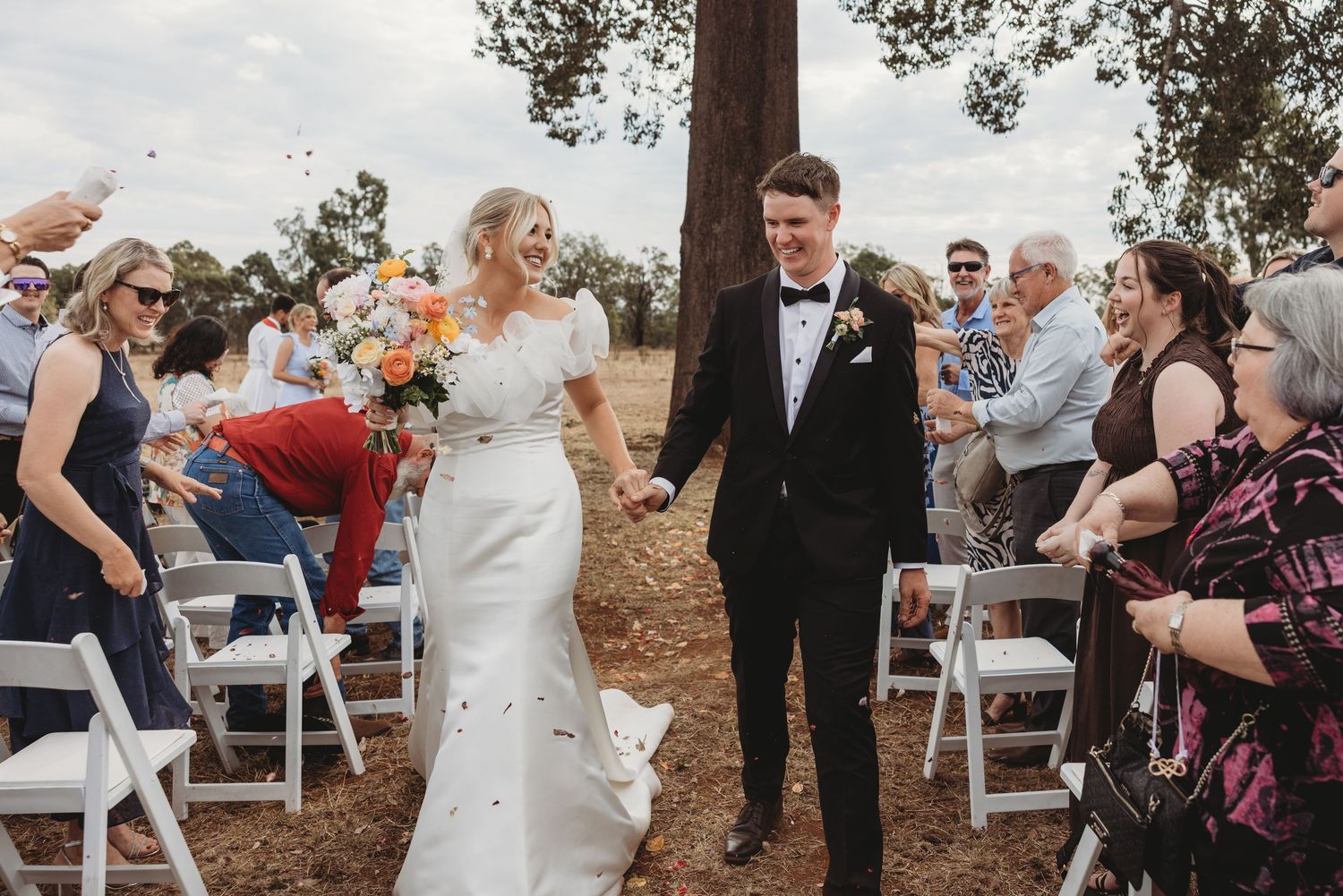 Wedding celebration with guests throwing confetti as couple walks down outdoor aisle.