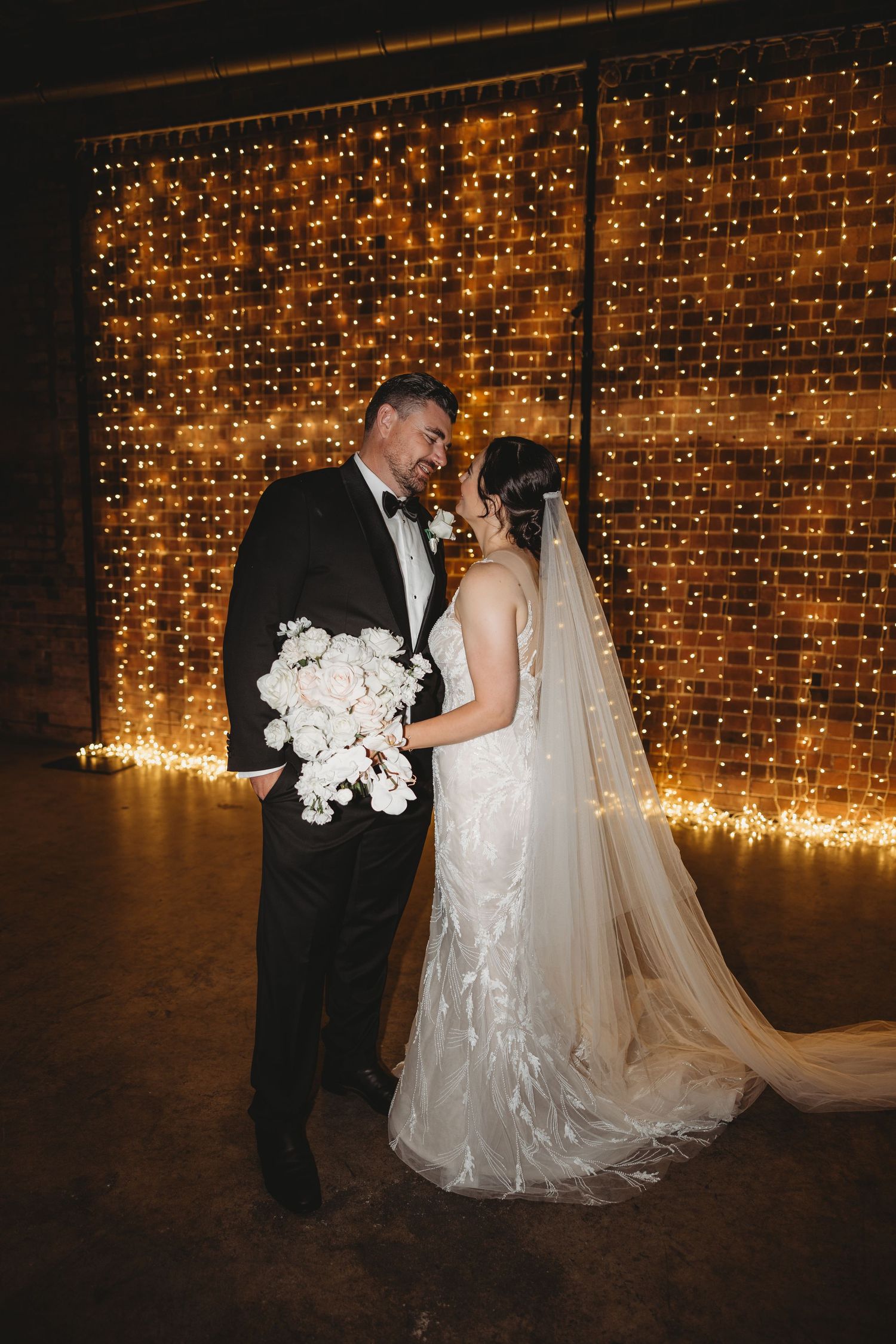 A romantic wedding couple shares a dance in front of a twinkling fairy light backdrop at an evening reception.